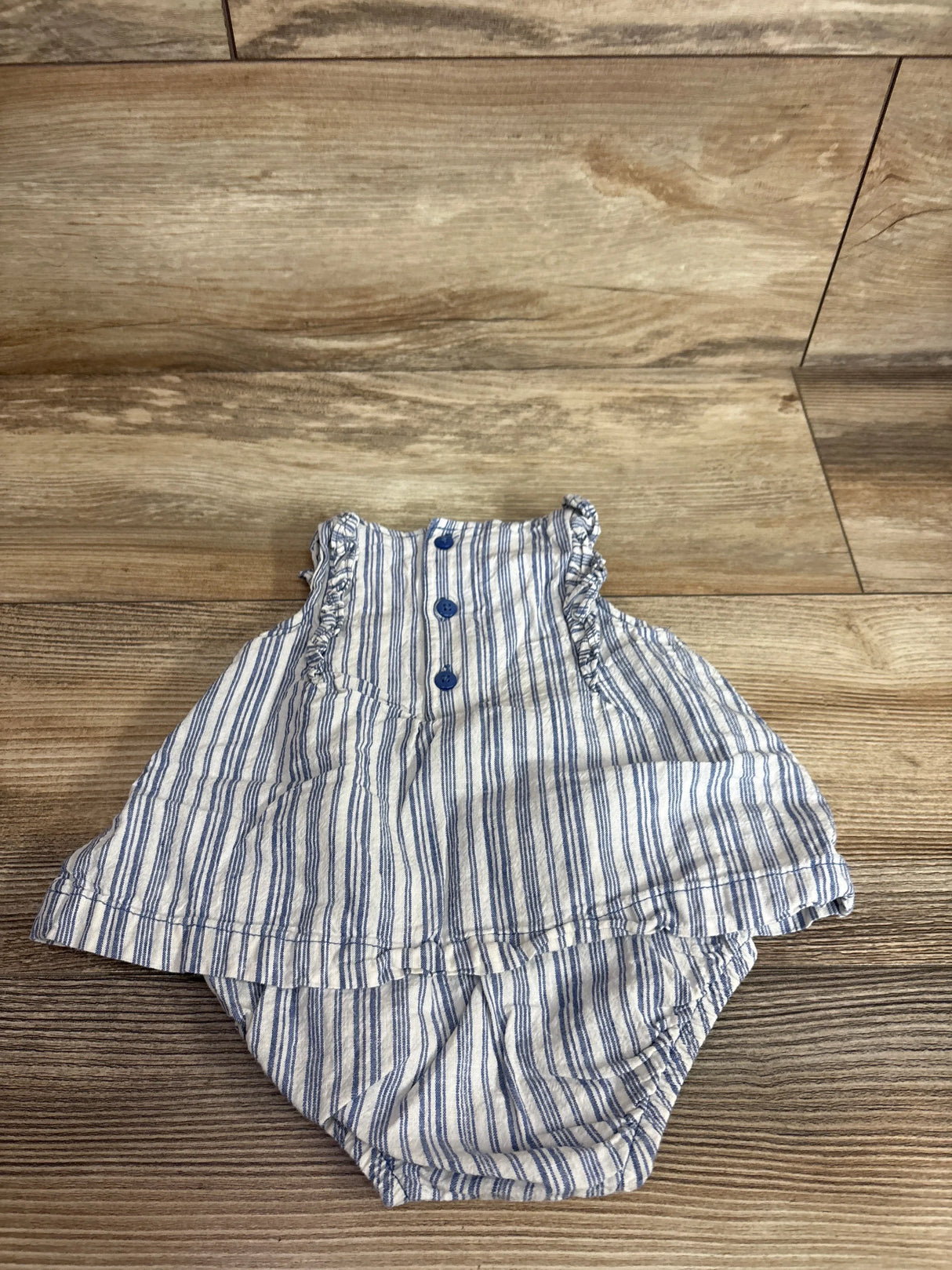 Top-down view of white and blue striped baby bodysuit dress size 3M showcasing ruffled shoulder straps, three blue front buttons, and attached bloomer on wood floor background
