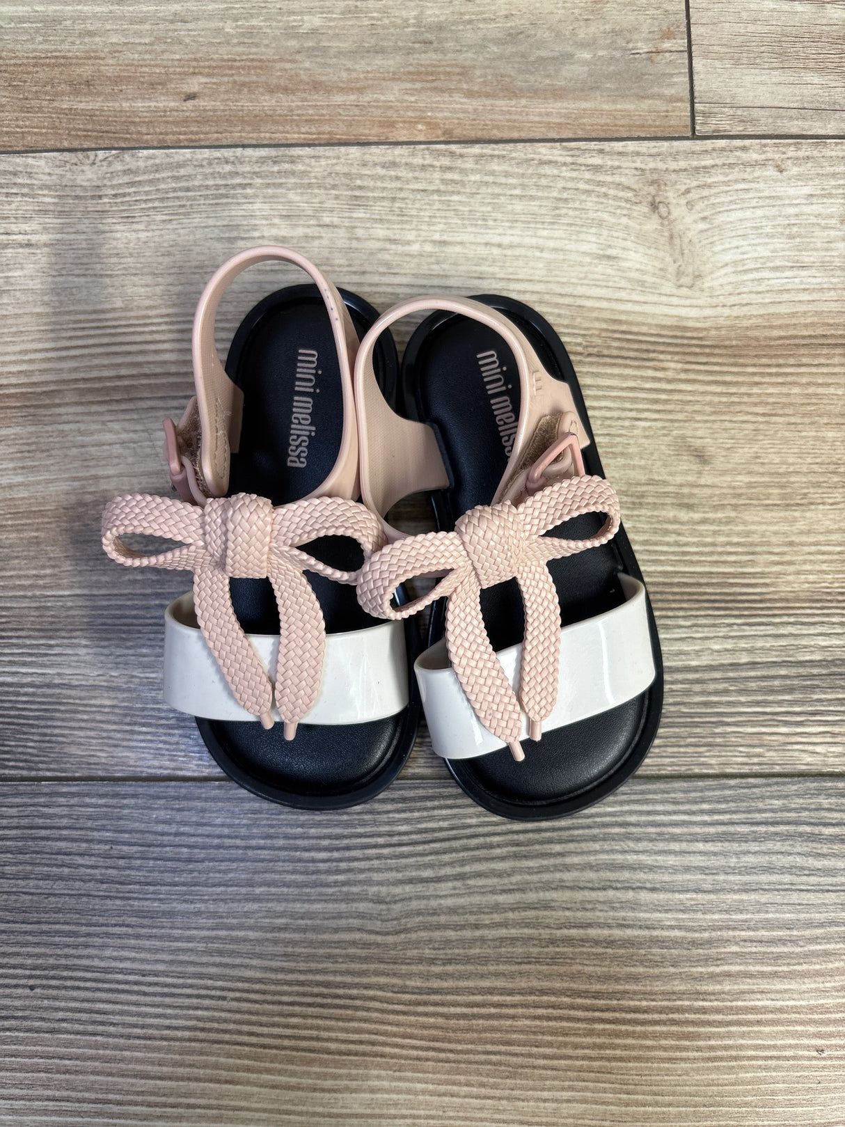 Top down view of Mini Melissa Mar sandals showing black footbed, white toe strap and pale pink braided bow on wood floor