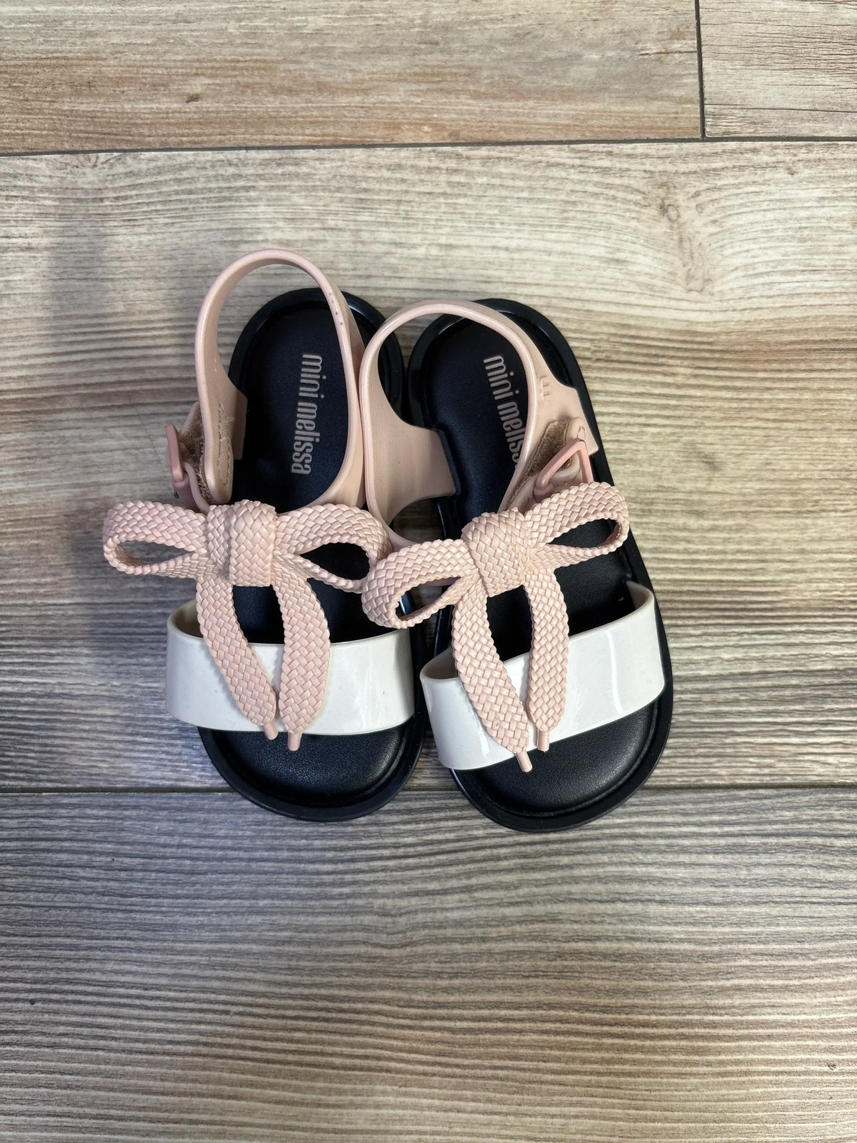 Top down view of Mini Melissa Mar sandals showing black footbed, white toe strap and pale pink braided bow on wood floor