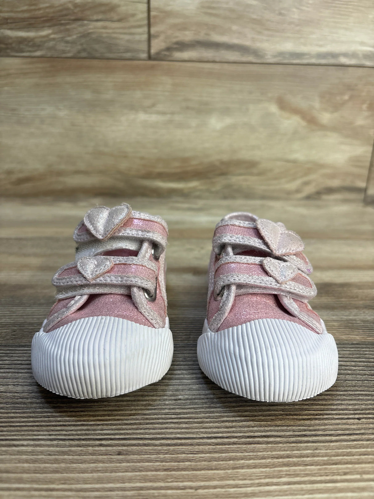 Pink toddler shoes with glitter heart straps and white rubber toes on wood floor