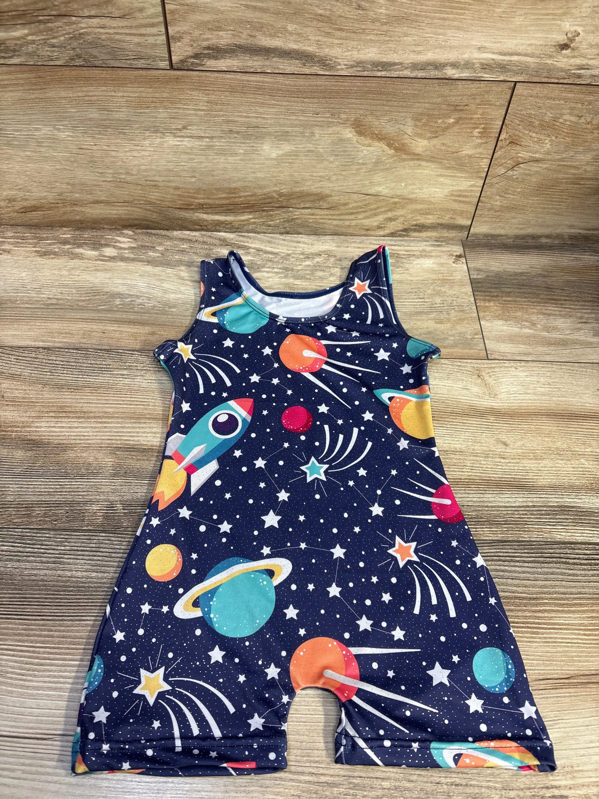 Flat lay of navy sleeveless toddler romper showcasing planets, rockets, and stars with wood floor background