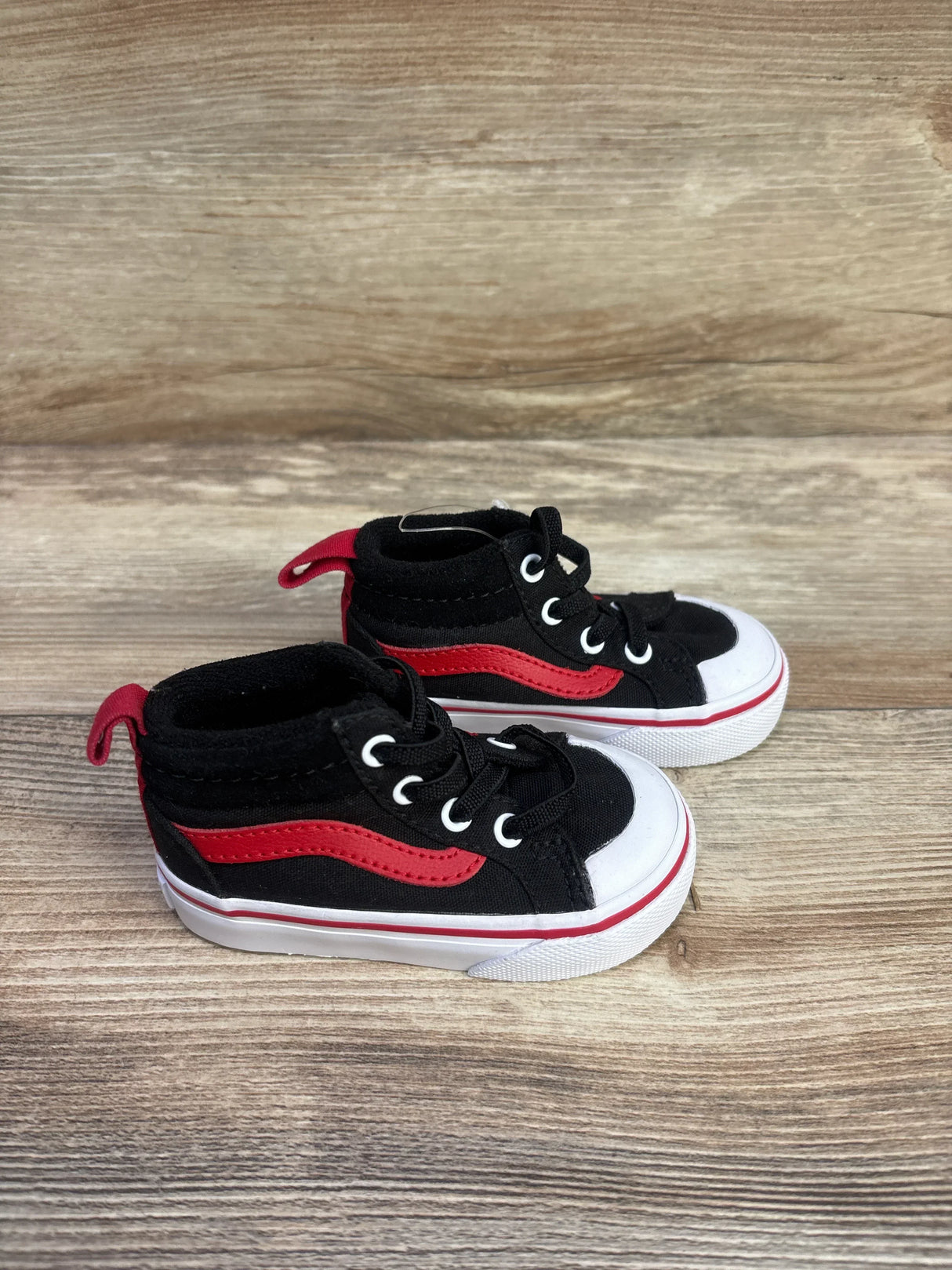 Three-quarter view of toddler black Vans mid-top sneakers with red stripe and white toe on wood background