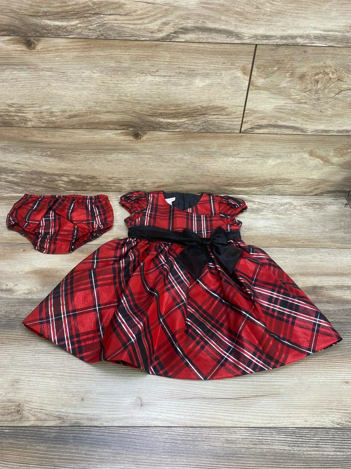 Flat lay of Bonnie Jean plaid toddler dress and bloomers showing puff sleeves, black waist bow on wood background