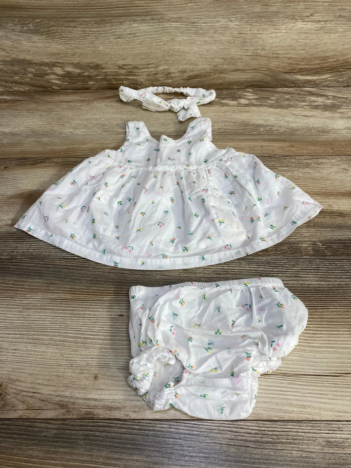 Front view of Baby Gap 3-piece white floral dress set with sleeveless dress, bloomers, and knotted headband on wood background
