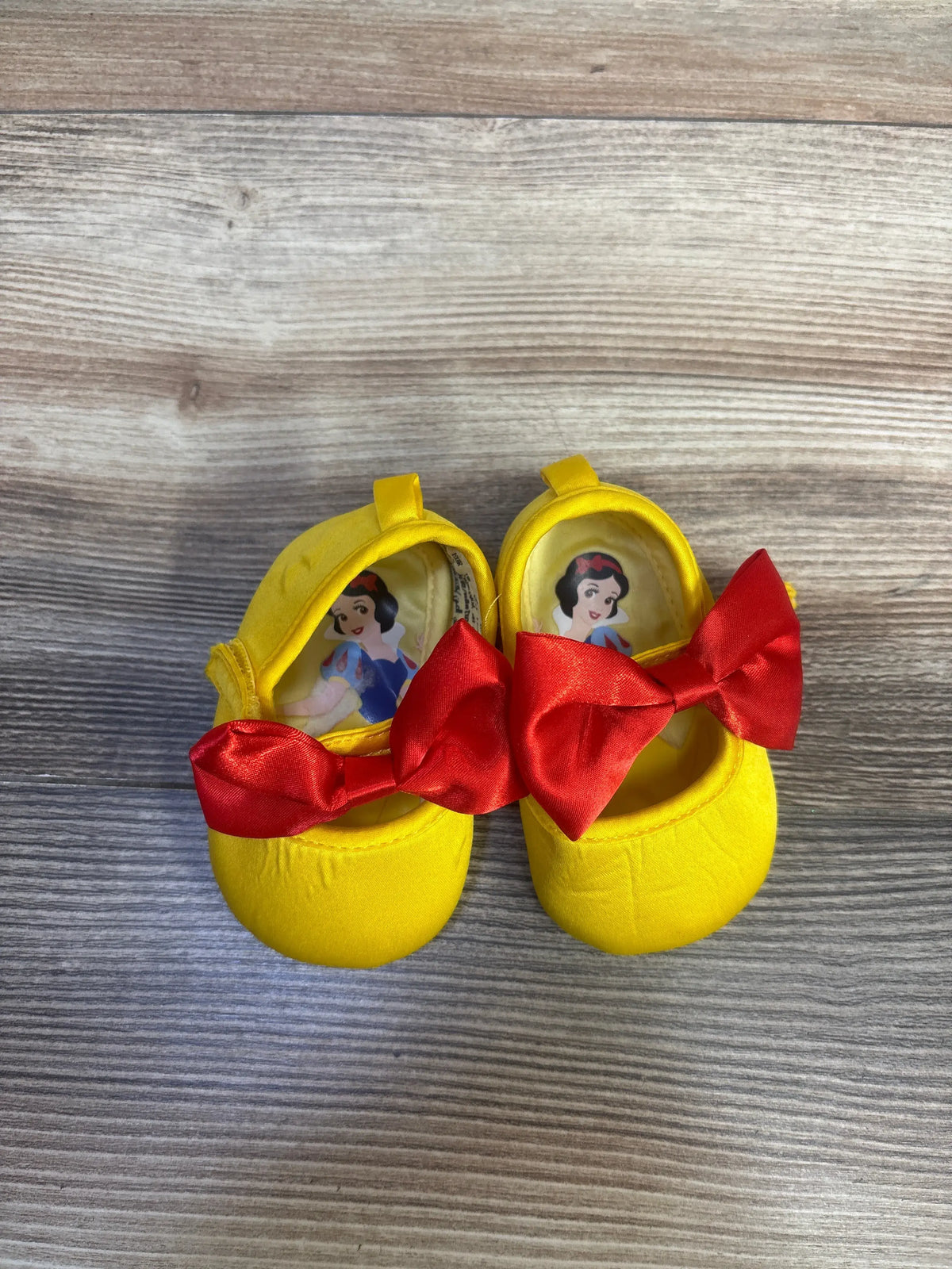 Top view of yellow Disney Baby Snow White soft sole shoes showcasing red bows and character insoles on a wood-grain background