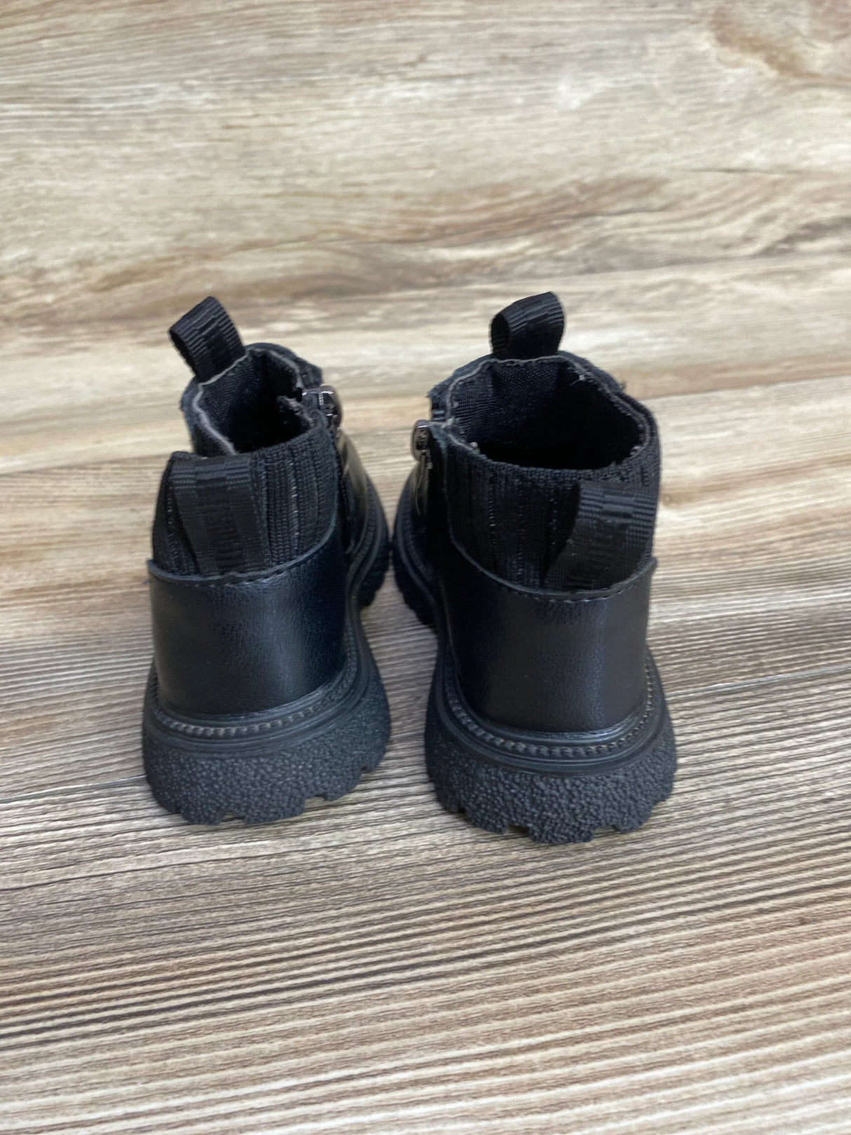 Back view of black toddler ankle boots showcasing side zipper closure heel pull tab and rugged lug sole on wood plank floor
