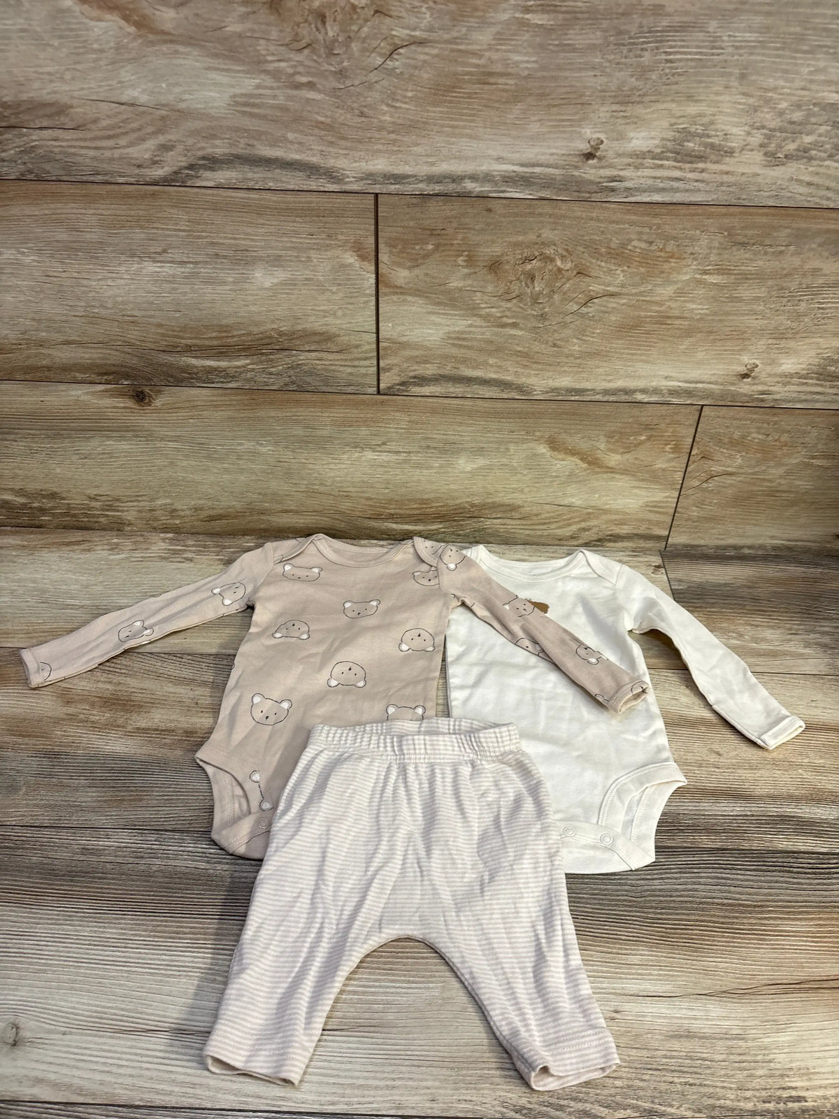 Flat lay of 3-piece long-sleeve bodysuit set (3M) with tan bear-print and white bodysuits plus beige striped pants on wood background