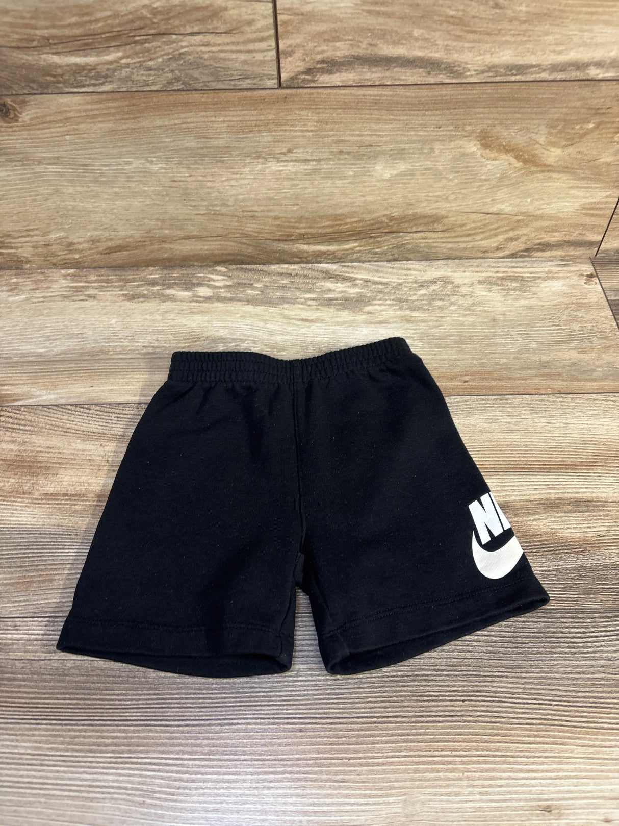 Flat lay of black Nike toddler shorts showing elastic waistband and white swoosh logo on wood floor background