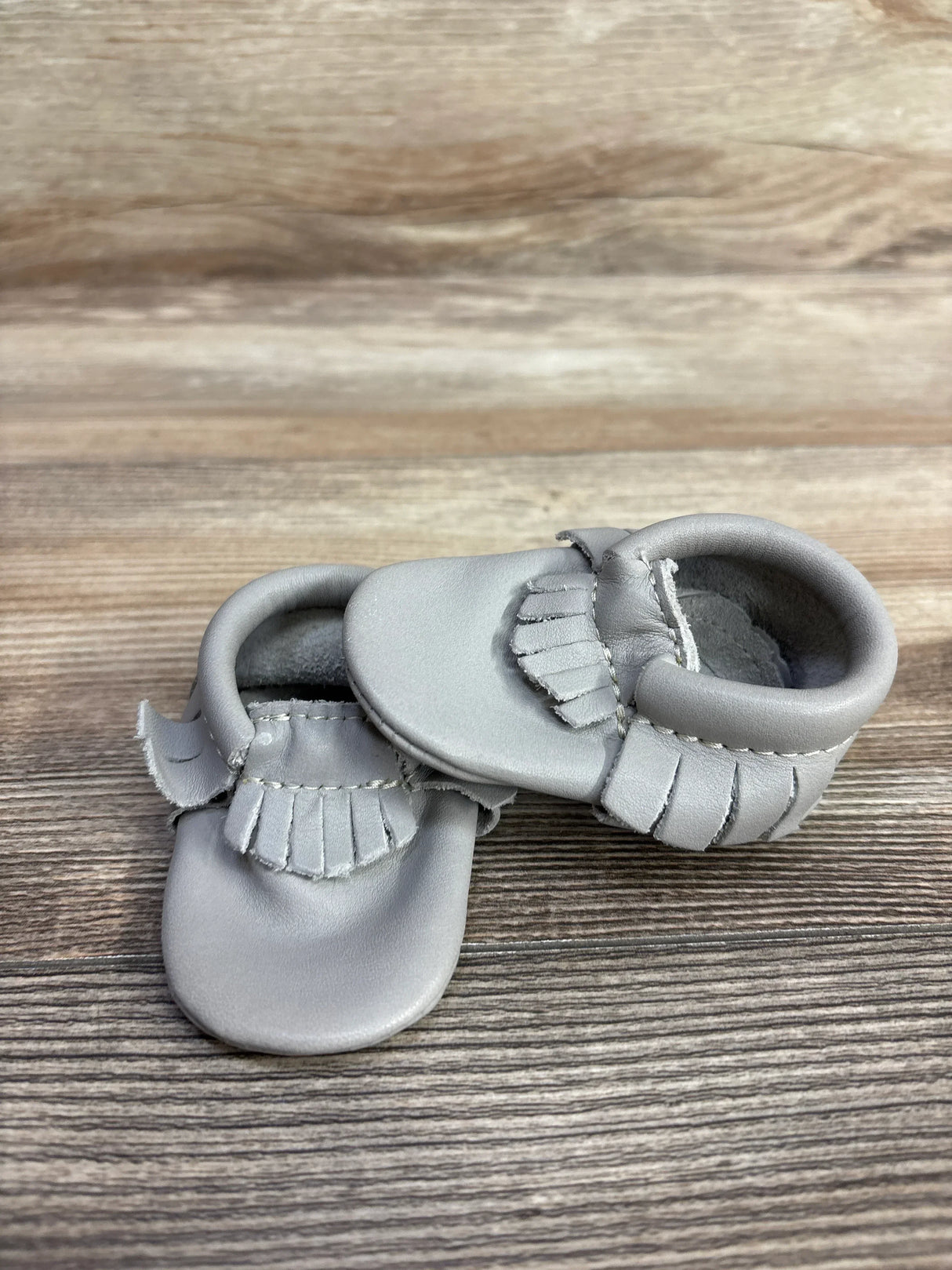 Pair of gray leather baby moccasins with fringe detail on wooden surface