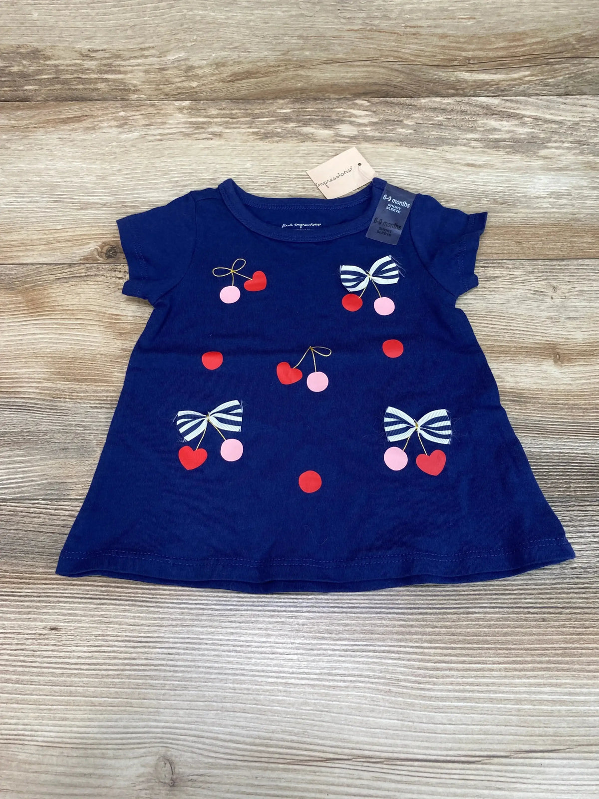 Flat lay of NEW First Impressions navy short sleeve shirt size 6-9m with cherry and striped bow print and hangtag on wood plank floor