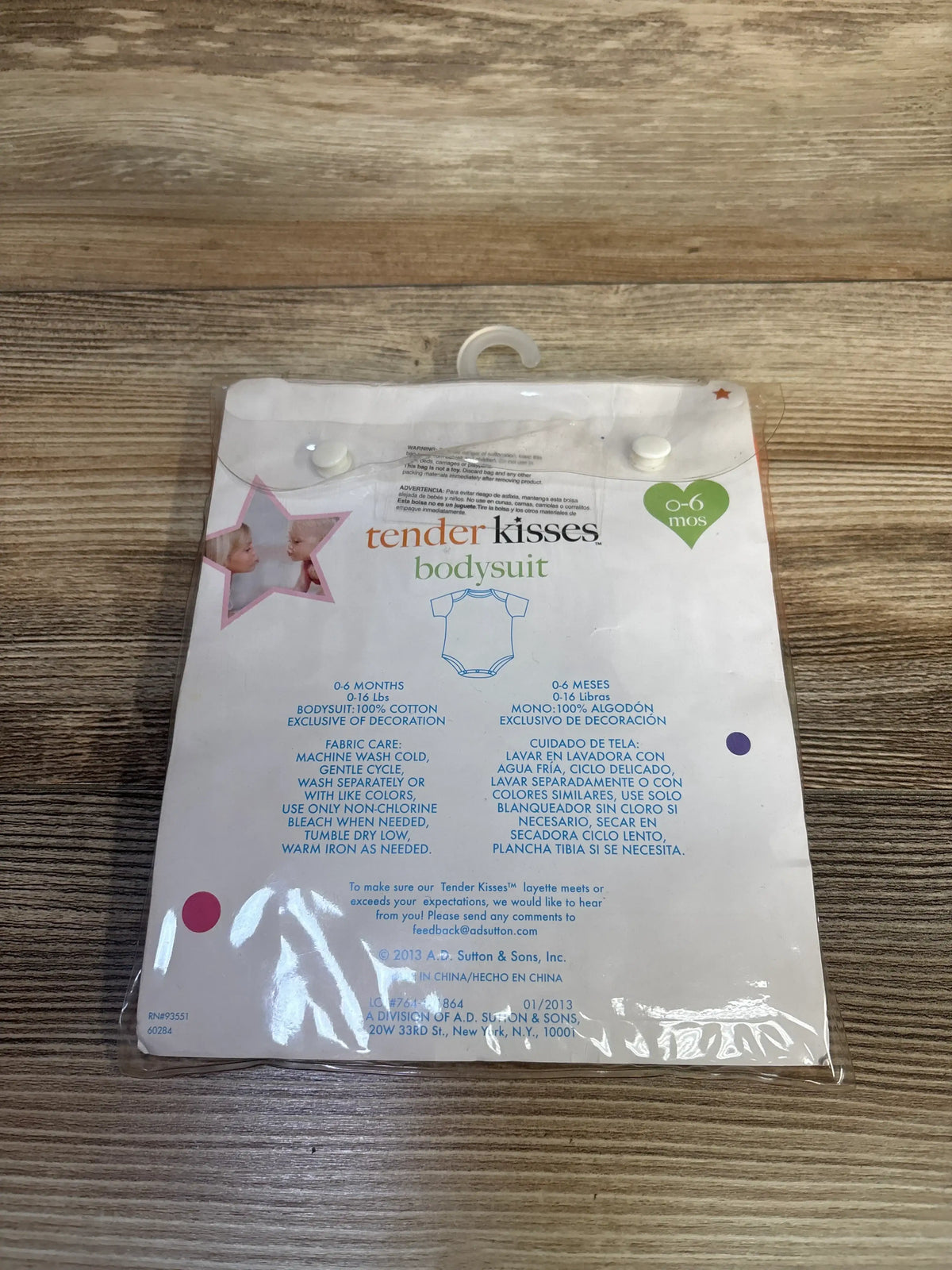 Back view of packaged Tender Kisses bodysuit showing 0 to 6 months sizing and cotton care details on a wood surface