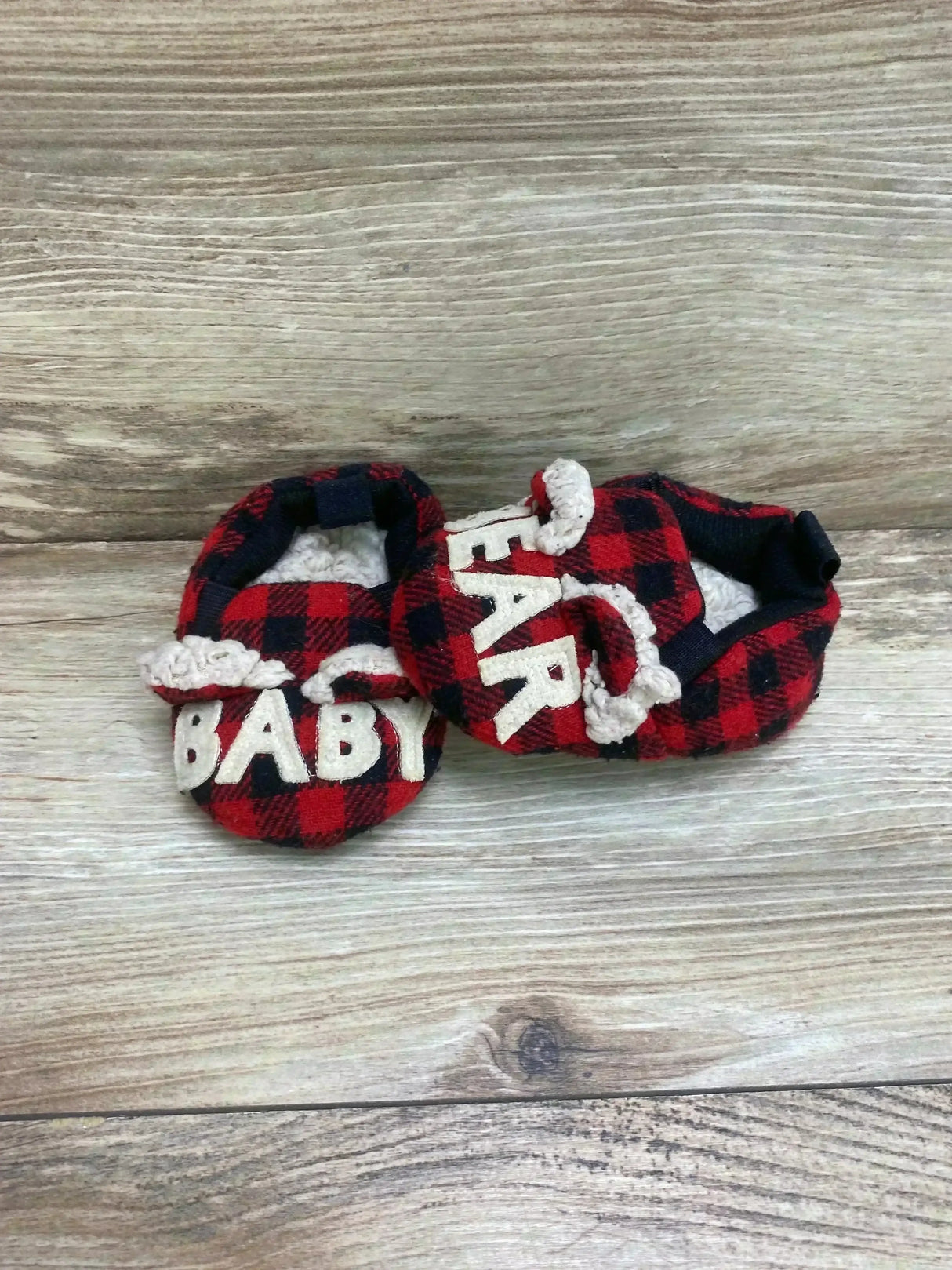 Front and side view of baby bear slippers in red black plaid with sherpa lining and white BABY letters on wood background