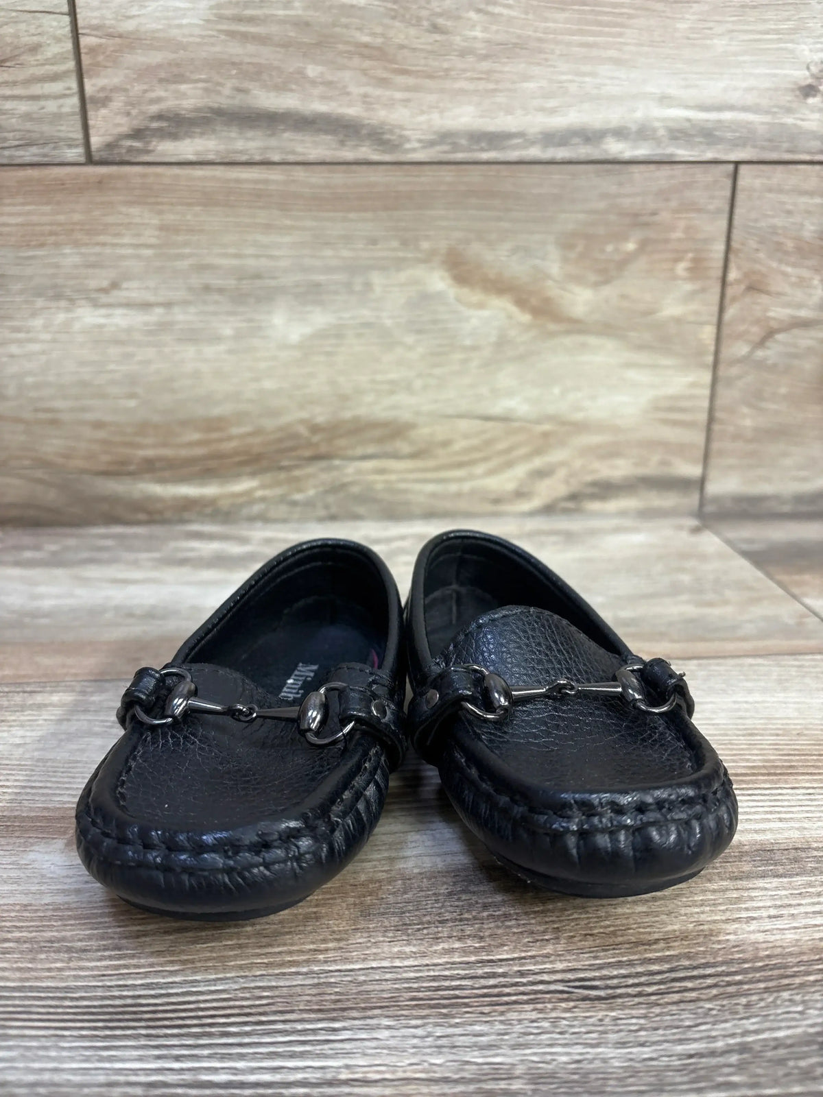 Front view of Minibella soft leather slip-on boat shoes, black size 6C, showing metal bit detail on wood floor