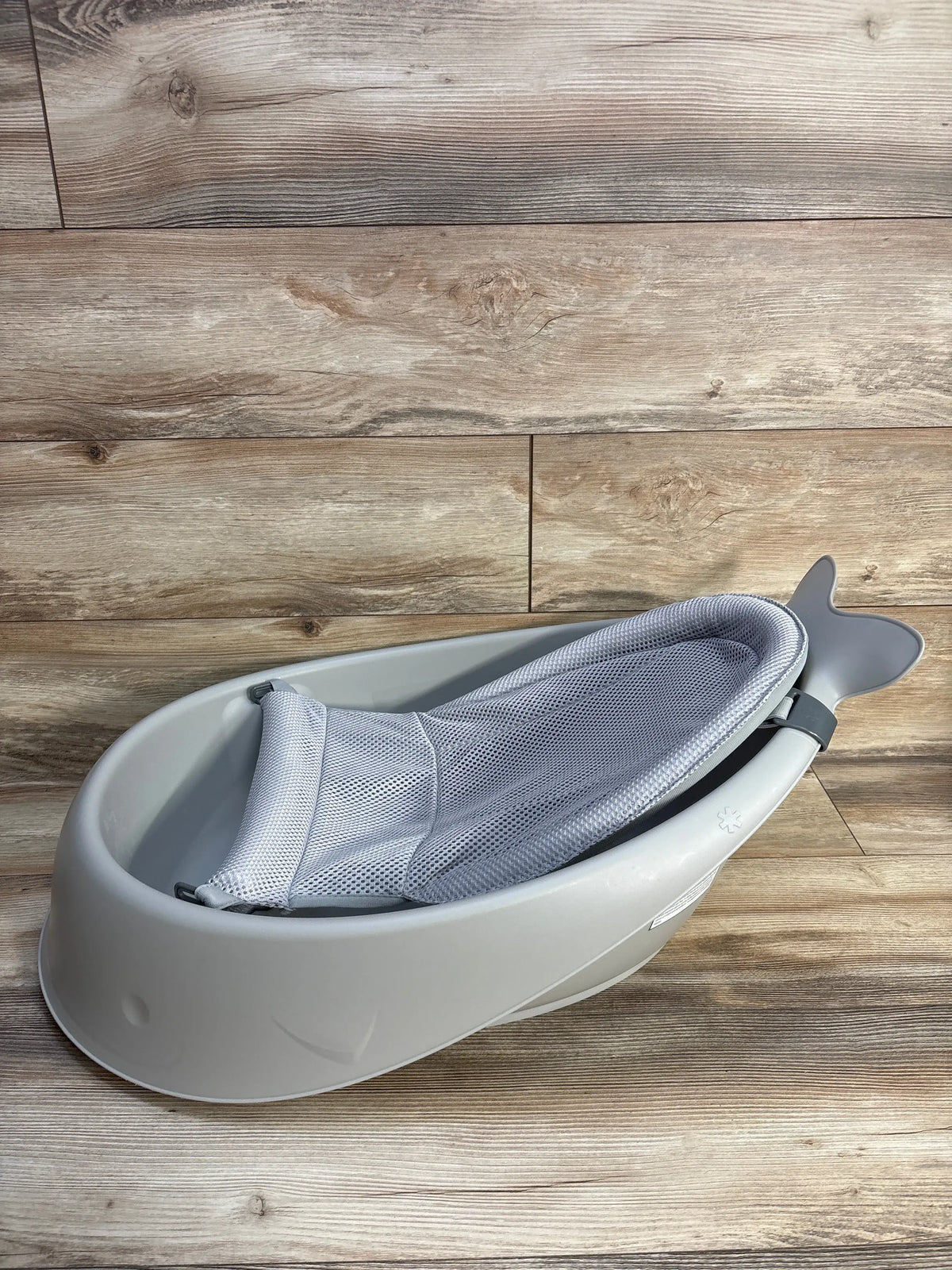 Angled view of grey Skip Hop Moby Smart Sling 3 in 1 baby bathtub showcasing its mesh Smart Sling on a wood floor