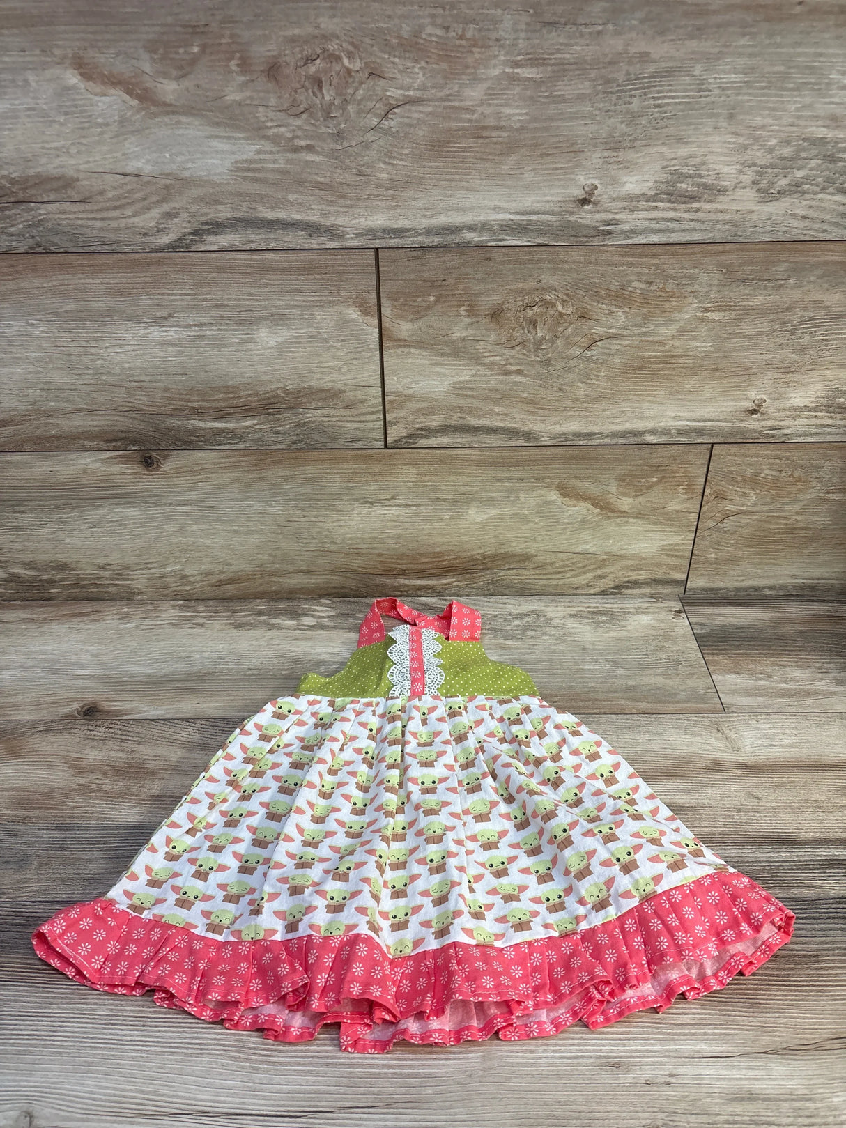 Flat lay of pink Baby Yoda toddler dress with green halter bodice, lace trim, and ruffled floral hem on wood floor