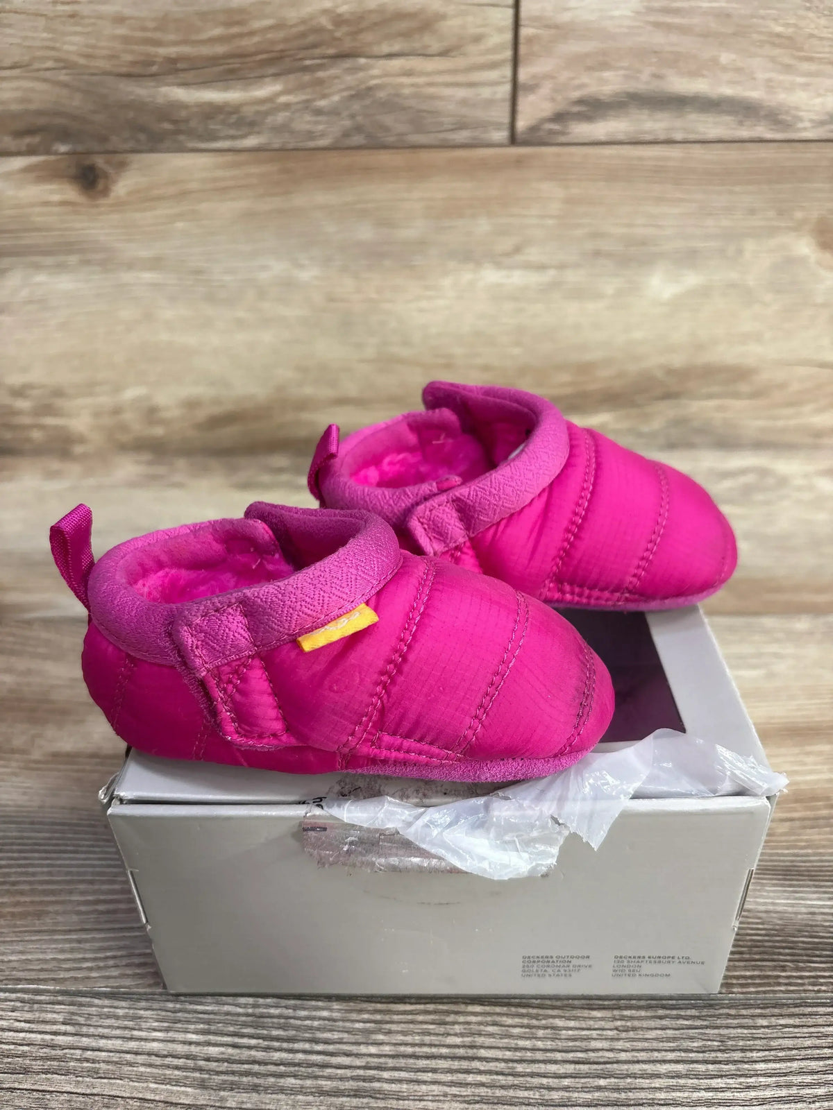 Pair of bright pink baby booties with soft lining on a worn white shoebox against wood background
