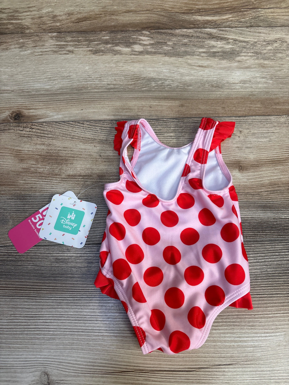 Pink and red polka dot baby swimsuit with ruffles, Disney Baby tag, on wood floor