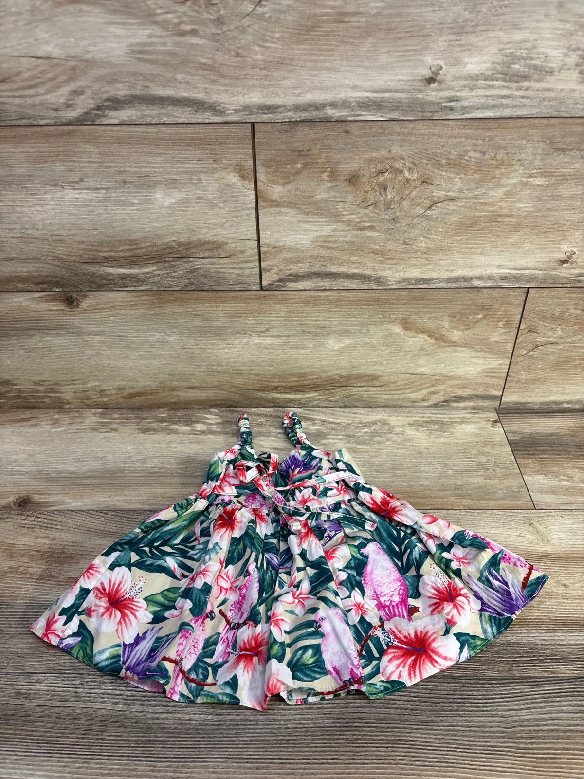 Flat lay of baby sleeveless dress showcasing tropical floral and pink parrot print with braided straps on light wood background