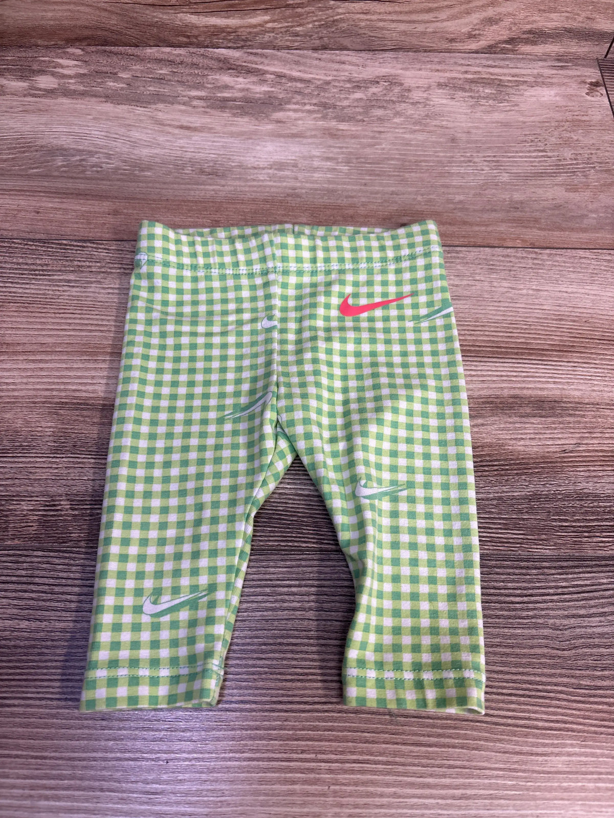 Front view of Nike green leggings showcasing a green and white checkered pattern and multiple Nike swoosh logos on wood flooring