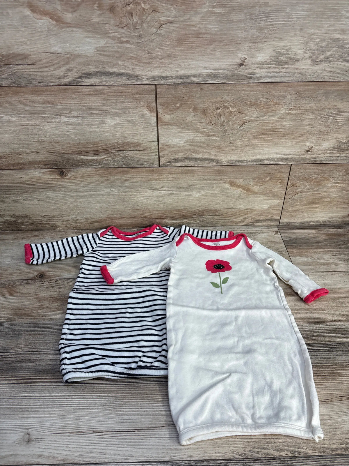 Top view of two cream Touched By Nature sleep gowns, one striped and one with a flower, both with pink trim on wood-look flooring