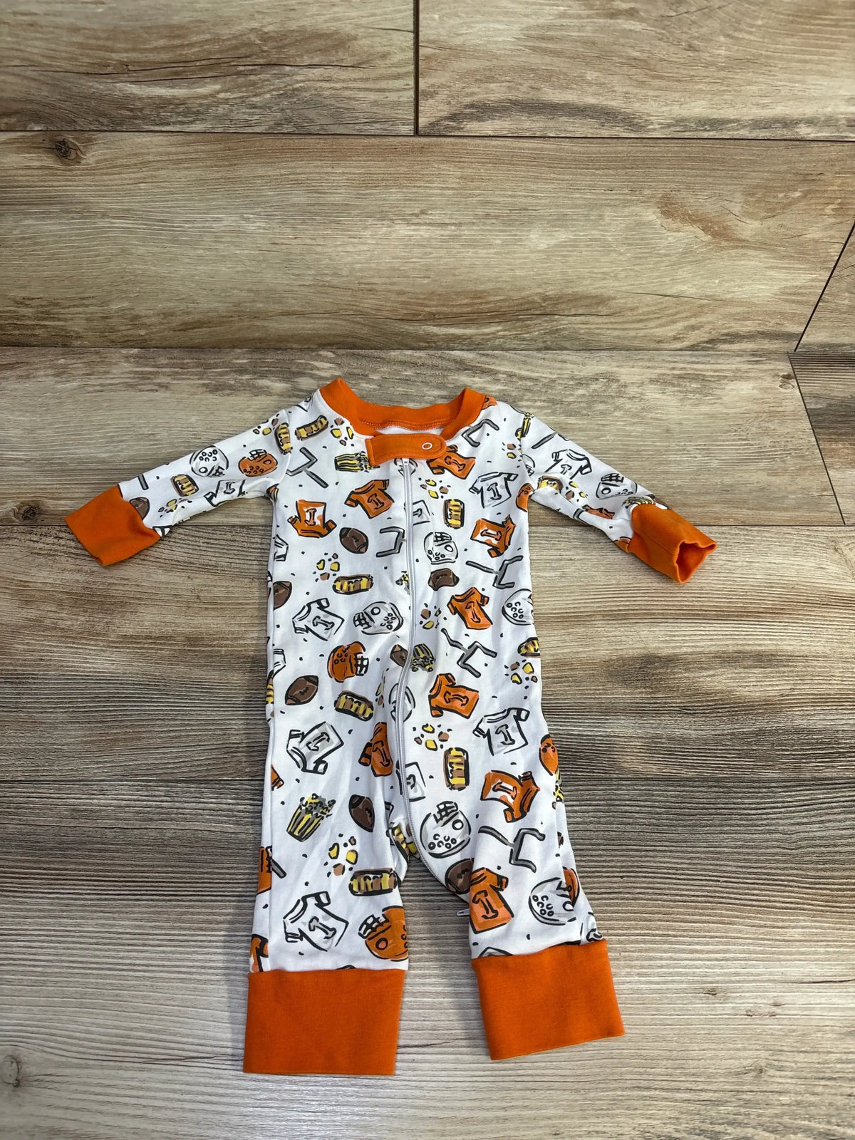 Top-down view of Magnolia Baby white footless sleeper with orange cuffs and football-themed print, front zipper on wood background