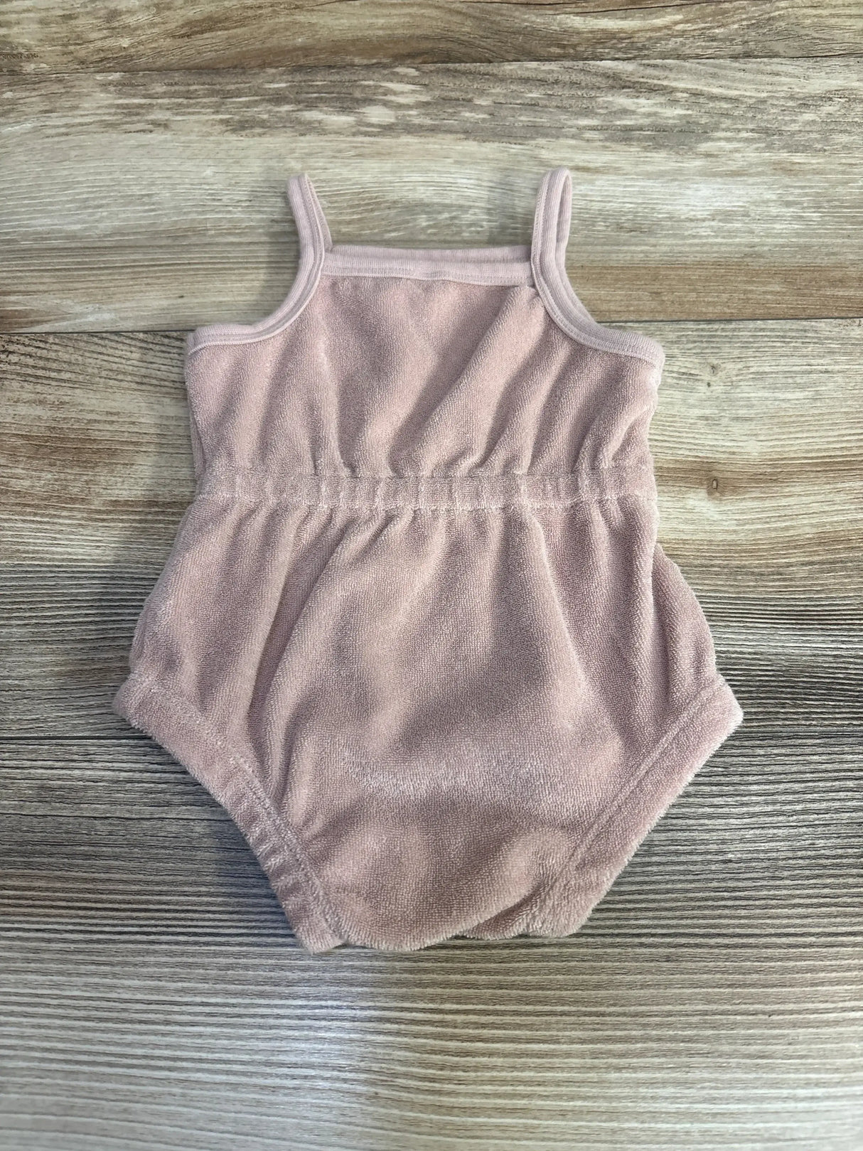 Flat lay of Quincy Mae pink terry romper with thin straps and elastic cinched waist on light wood background