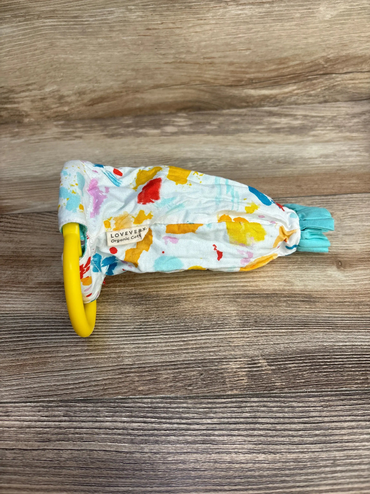 Top down view of Lovevery Organic Cotton Sensory Pouch showcasing multicolor splash print with yellow clip ring and tassel on wood plank floor