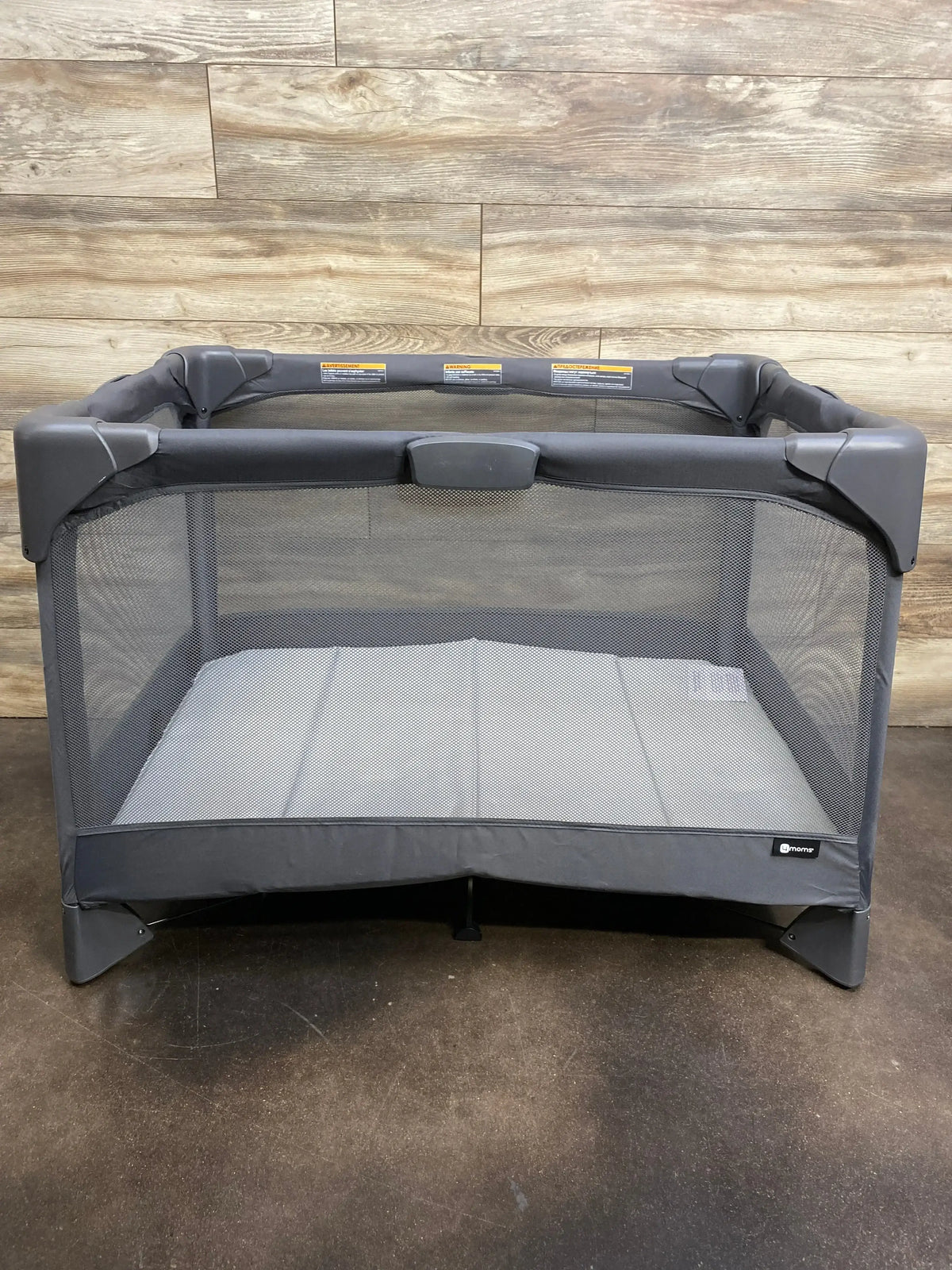 Front view of 4moms Breeze Go playard in grey showing mesh sides and padded mattress on wood-plank background