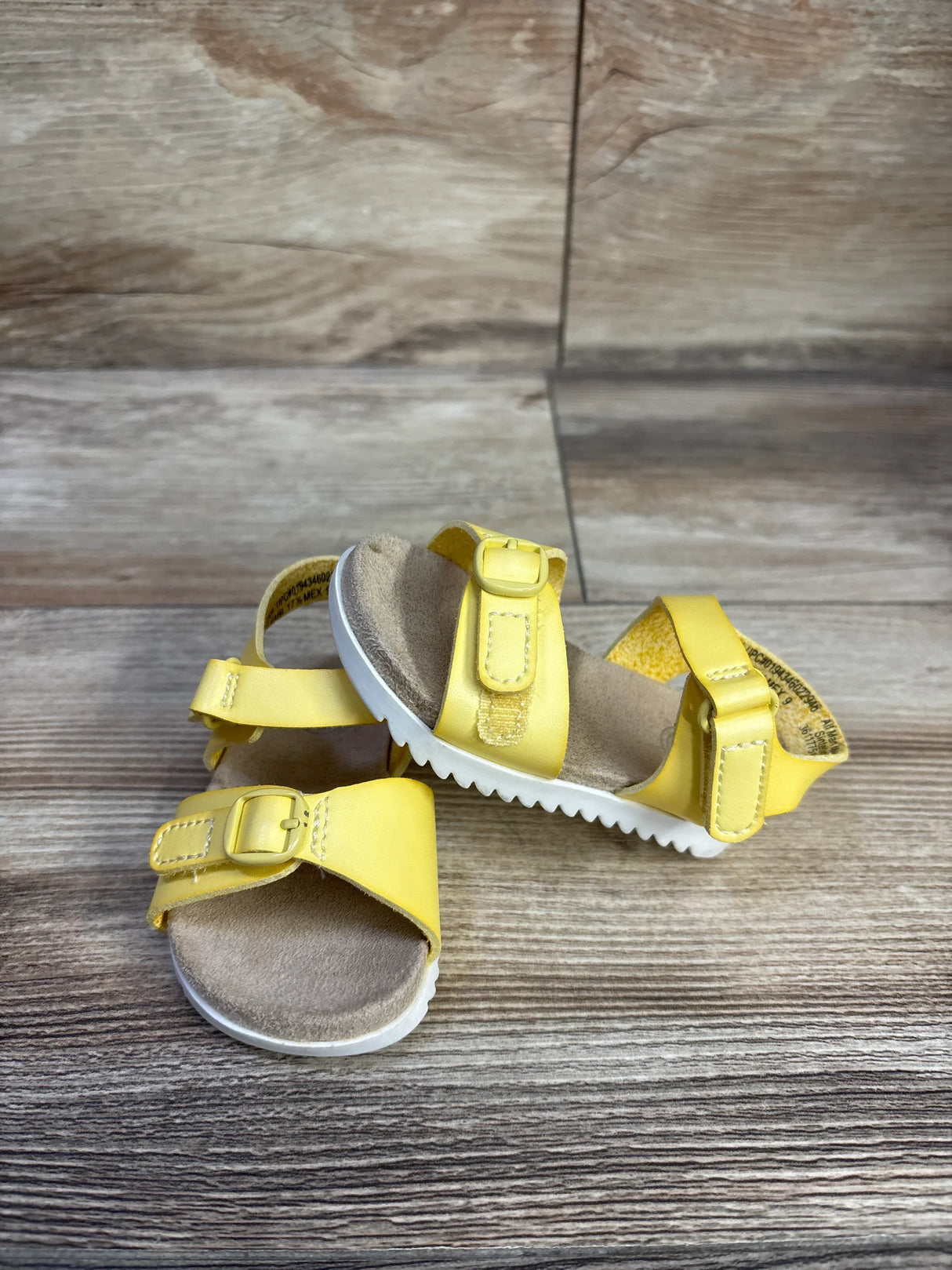 Three-quarter view of yellow toddler buckle sandals showing adjustable buckle straps, cushioned tan footbed and white ridged sole on wood background
