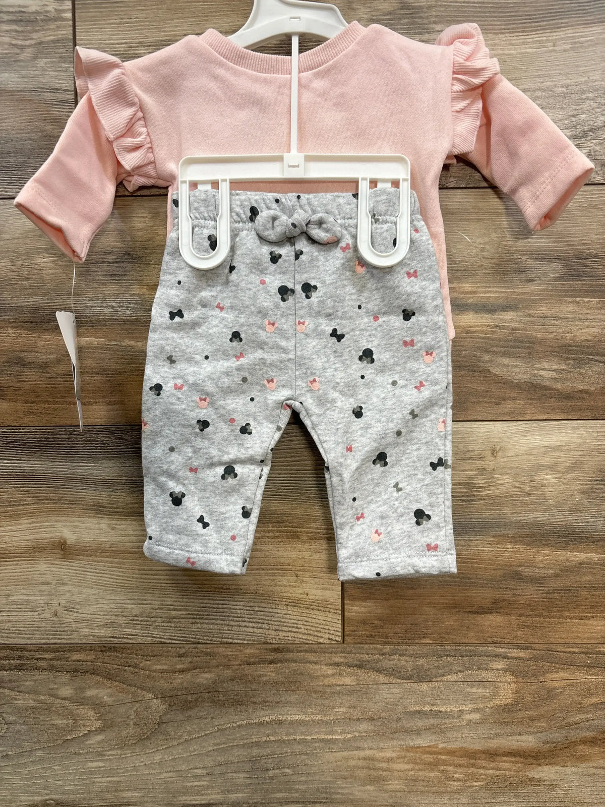 Pink long-sleeve baby top with ruffled shoulders and gray pants with black and pink mouse silhouette prints on wooden floor