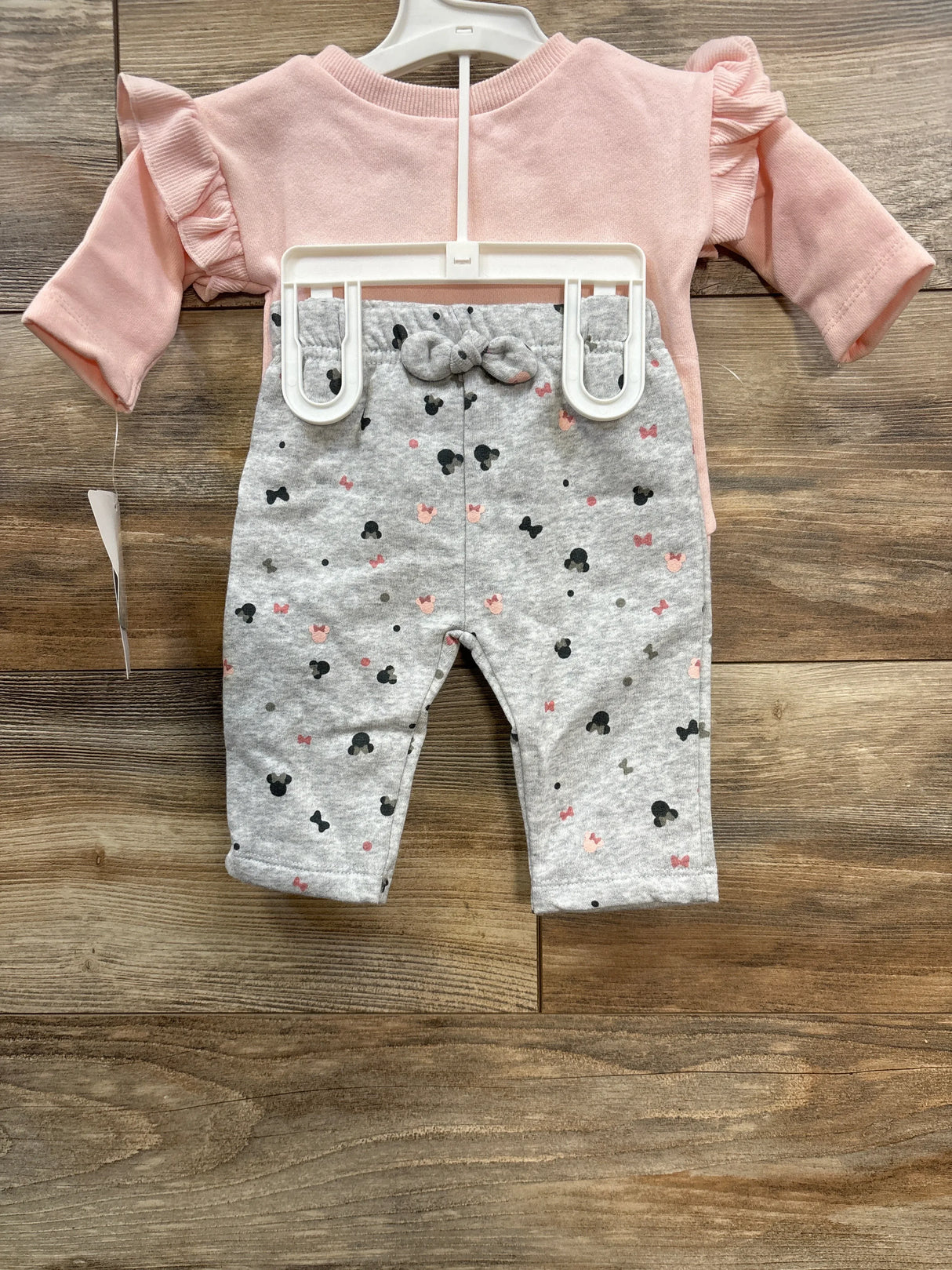 Pink long-sleeve baby top with ruffled shoulders and gray pants with black and pink mouse silhouette prints on wooden floor