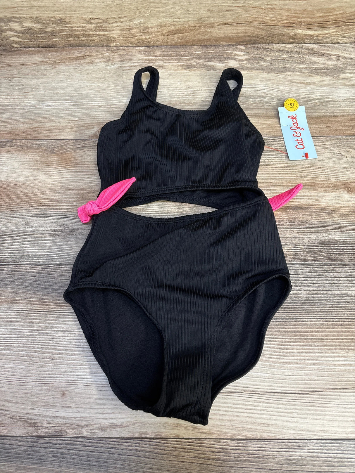 Top-down view of Cat & Jack black ribbed one-piece swimsuit with midriff cut-out and pink side ties on wood background