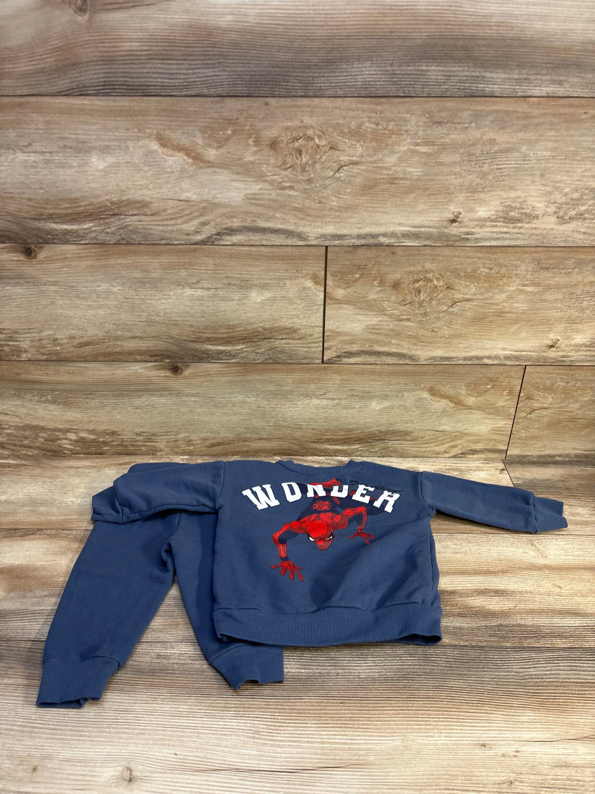 Flat lay of pullover sweatshirt and joggers set showcasing red Spider-Man graphic and WONDER text on navy sweatshirt with wood background
