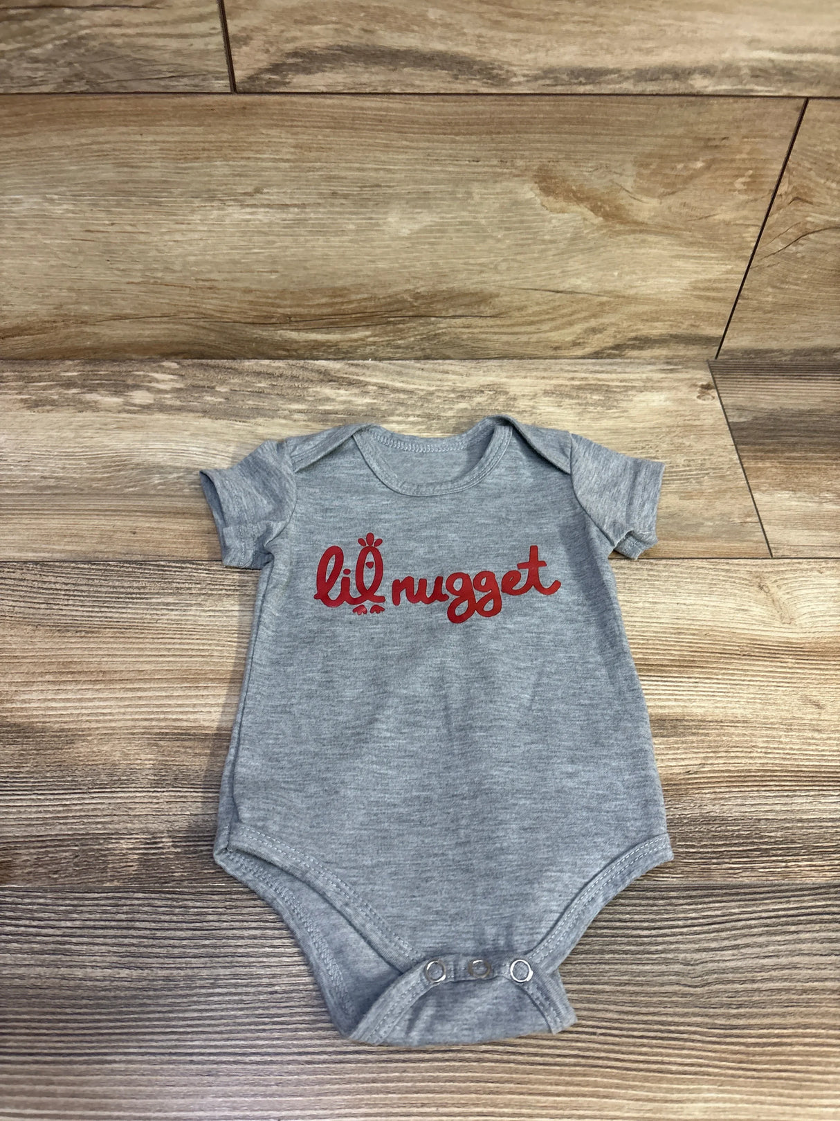 Flat lay of grey Sprout Joy Lil Nugget baby bodysuit showcasing red lil nugget chest print on wood plank background