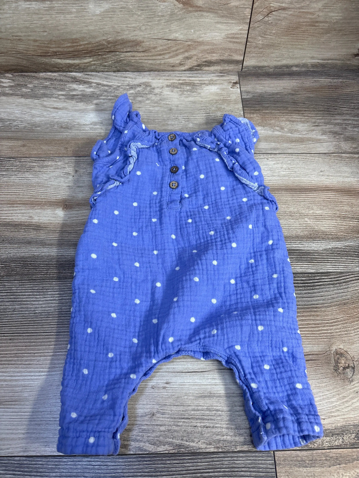 Flat lay of purple gauze baby jumpsuit with white polka dots, ruffle sleeves and three front wooden buttons on light wood background