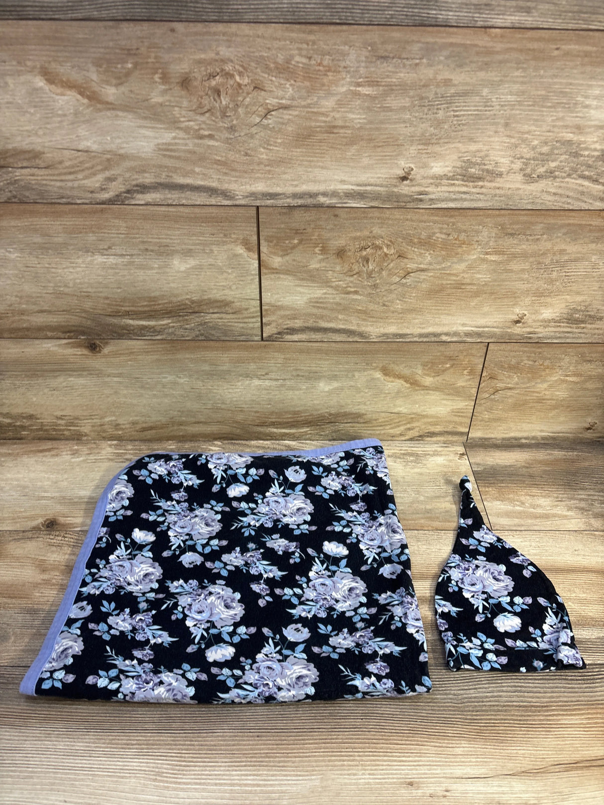 Top-down view of 2-piece floral swaddle and matching hat with lavender roses on black fabric, wood background
