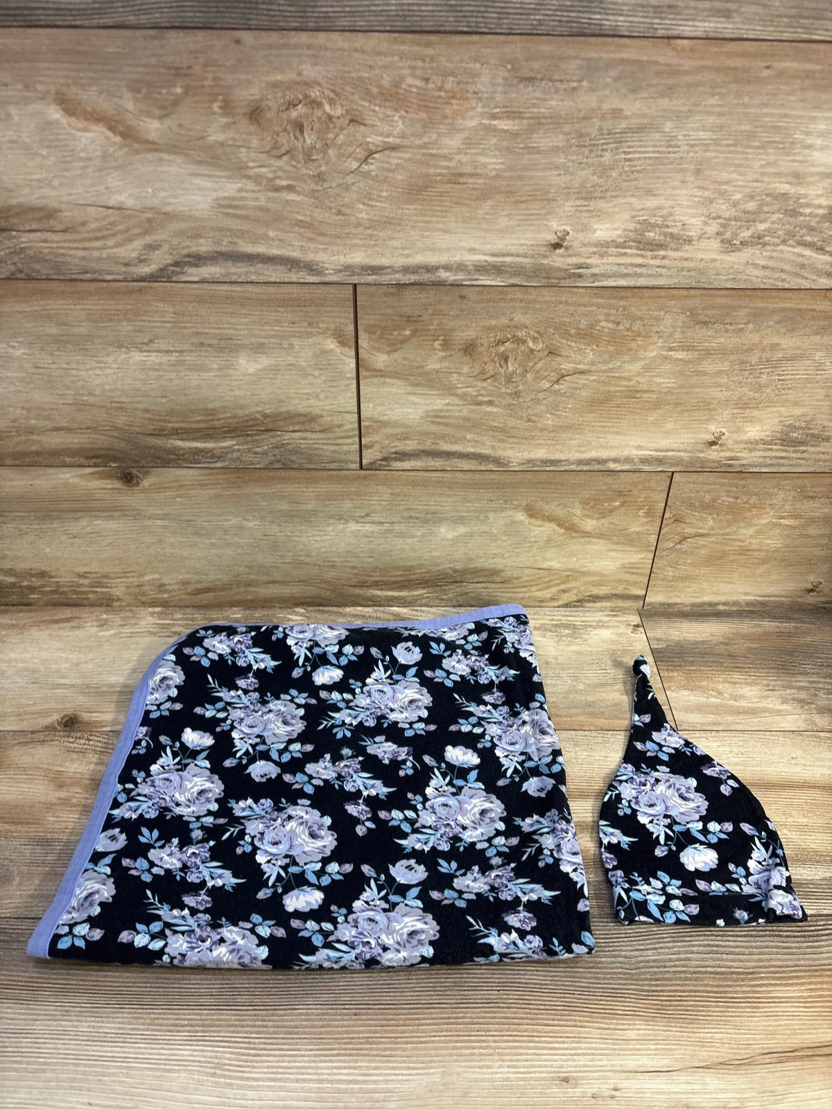 Top-down view of 2-piece floral swaddle and matching hat with lavender roses on black fabric, wood background