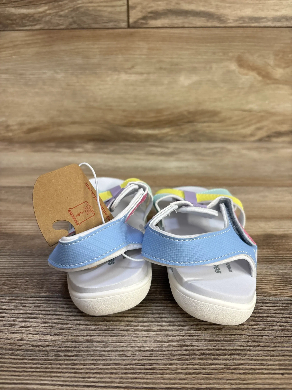 Front view of See Kai Run Shayna toddler sandals showcasing blue front strap and pastel multicolor ankle straps with Velcro closure on wood floor