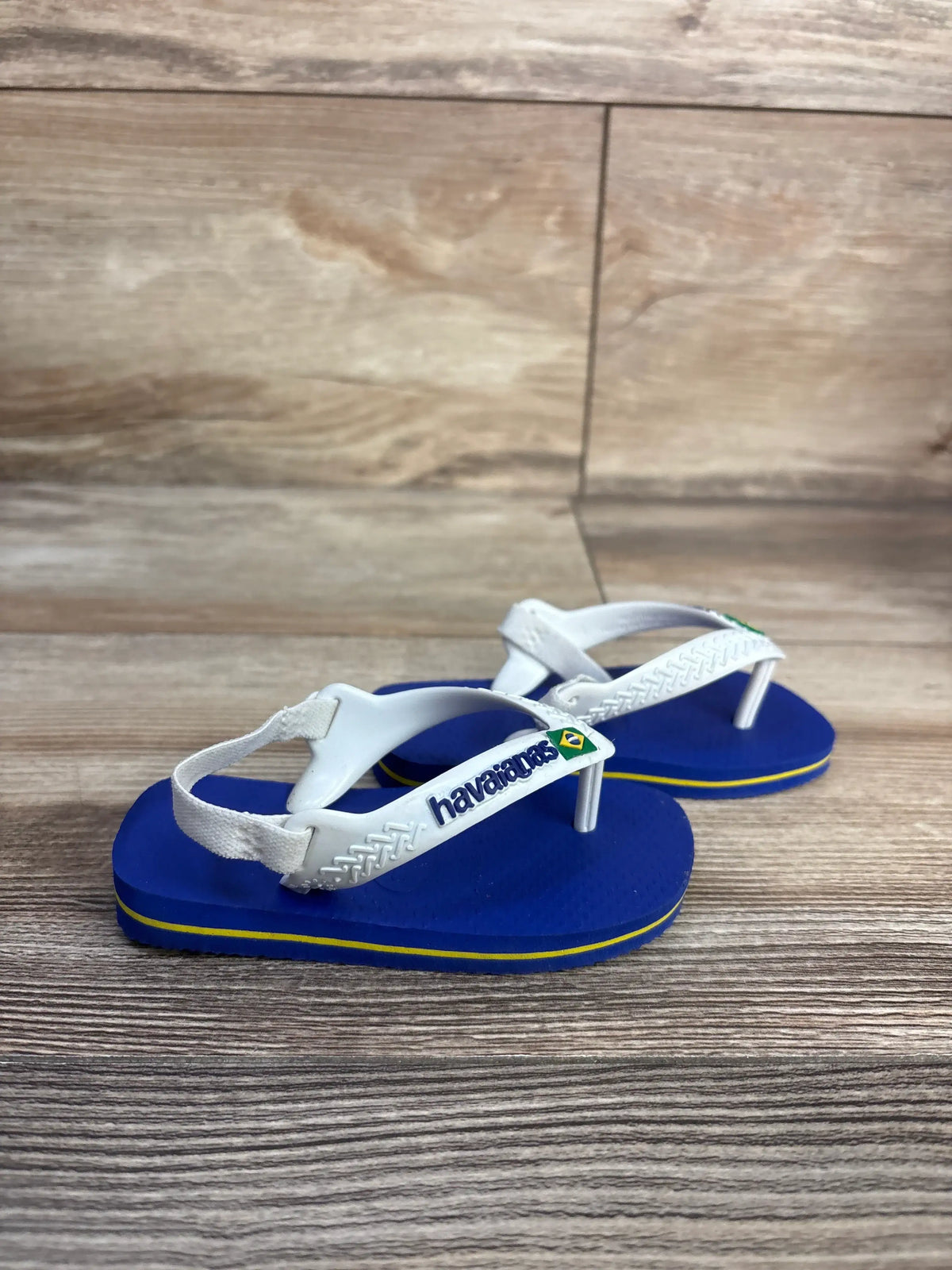 Front three-quarter view of Havaianas flip flops showcasing blue soles, white thong straps, and elastic ankle straps on a wood-grain background