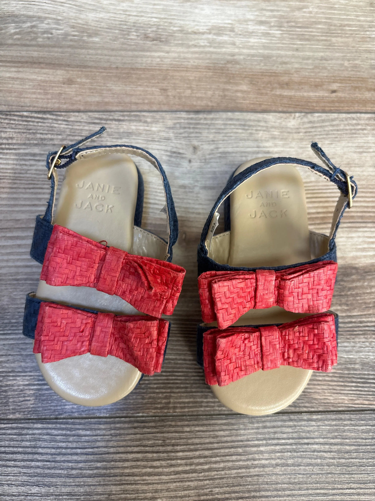 Top view of Janie and Jack toddler sandals with navy strap and red woven raffia bow on wood plank floor
