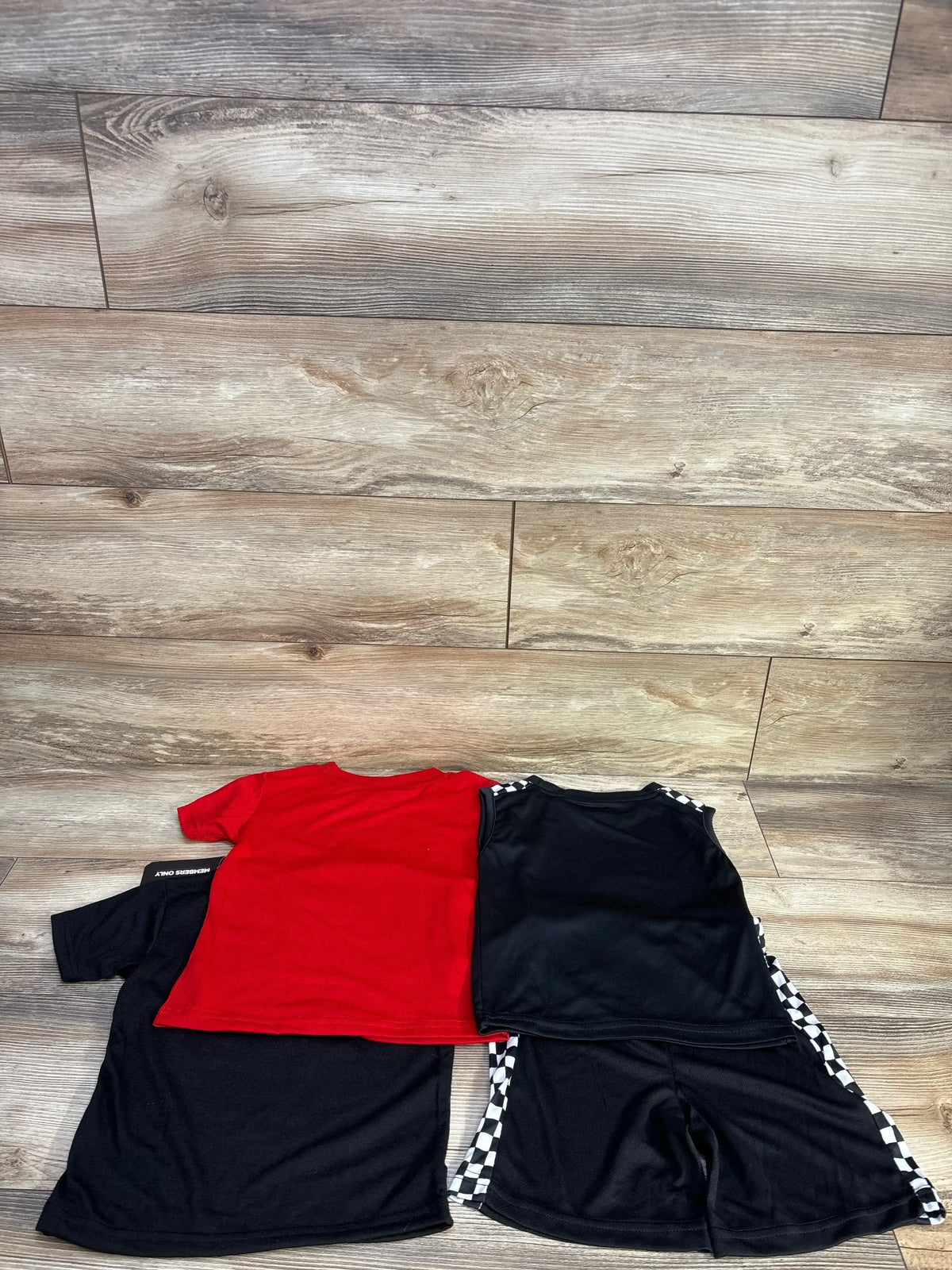 Flat lay of Member’s Only 4-piece toddler set size 4T: red tee, black tank, black shorts with checkered side panels on wood background