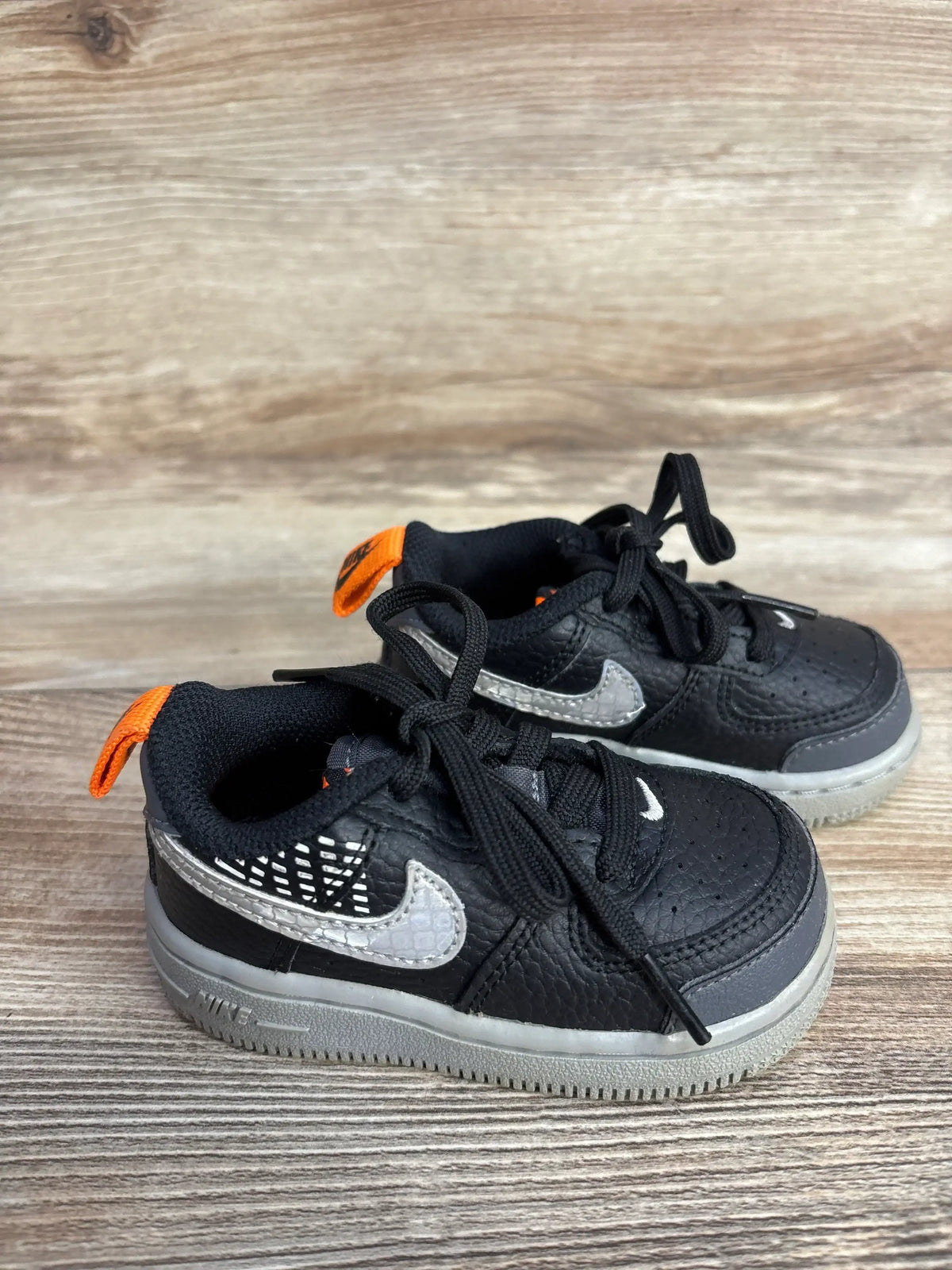 Three-quarter view of toddler Nike Air Force 1 LV8 2 TD sneakers showcasing black leather upper, silver textured Swoosh and orange heel tab on wood floor