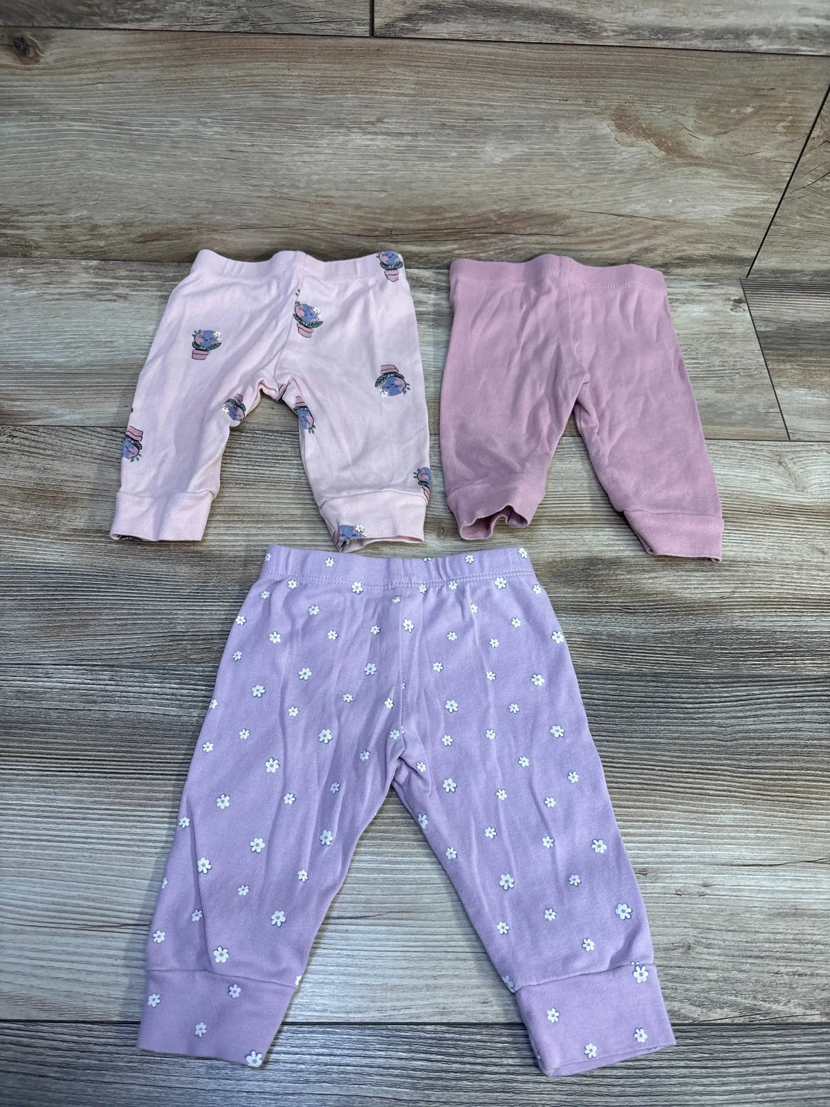 Top view of Pekkle three-pack pink pants in size 3 months, with floral and printed designs on a wood-look floor