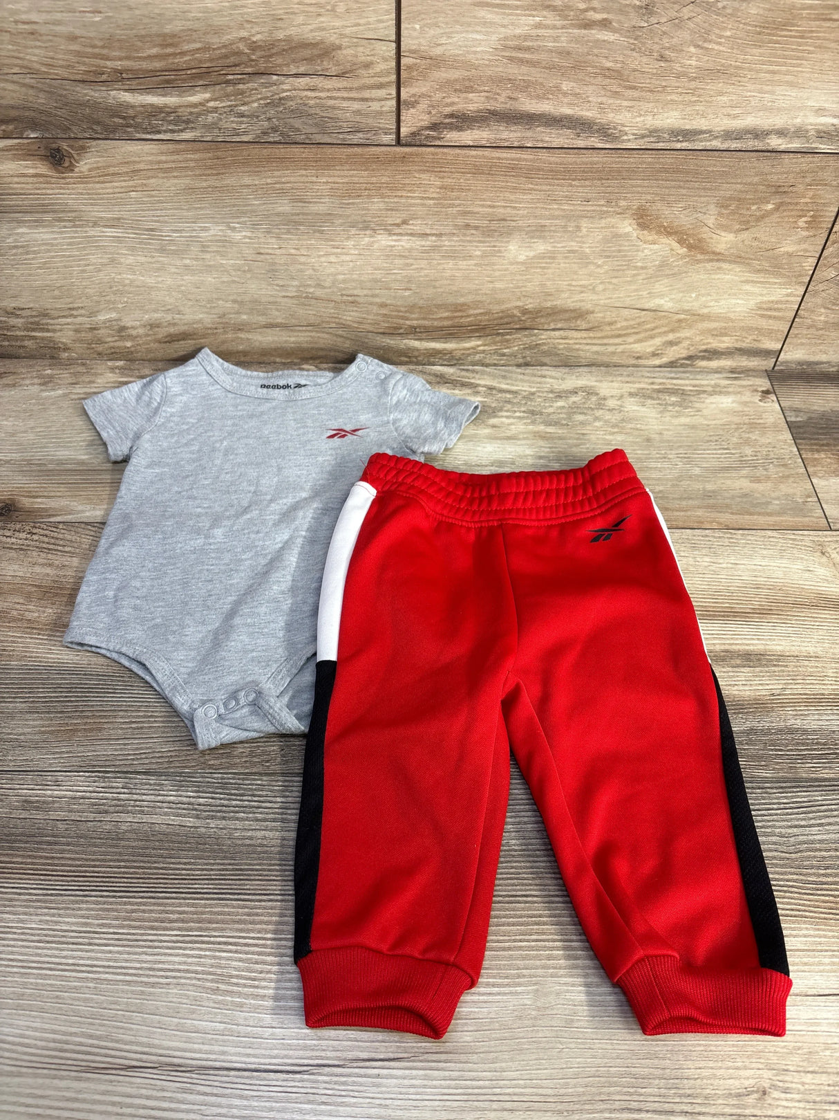 Top-down view of Reebok 2-piece infant set: grey bodysuit with red logo and red joggers with black side panels on wood floor