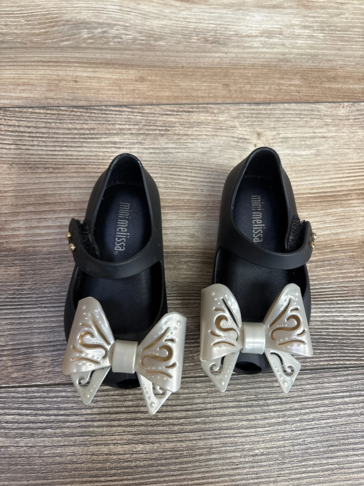 Pair of black Mini Melissa children's shoes with large decorative cream bows on wooden floor