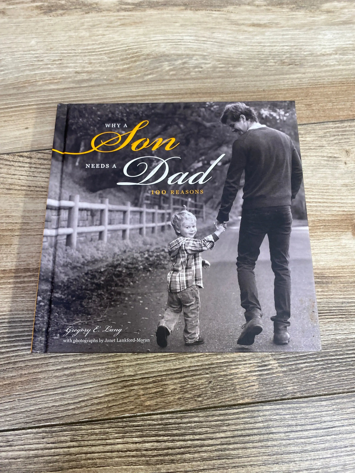 Overhead view of Why a Son Needs a Dad hardcover showing black-and-white cover of father holding toddler's hand on wood floor