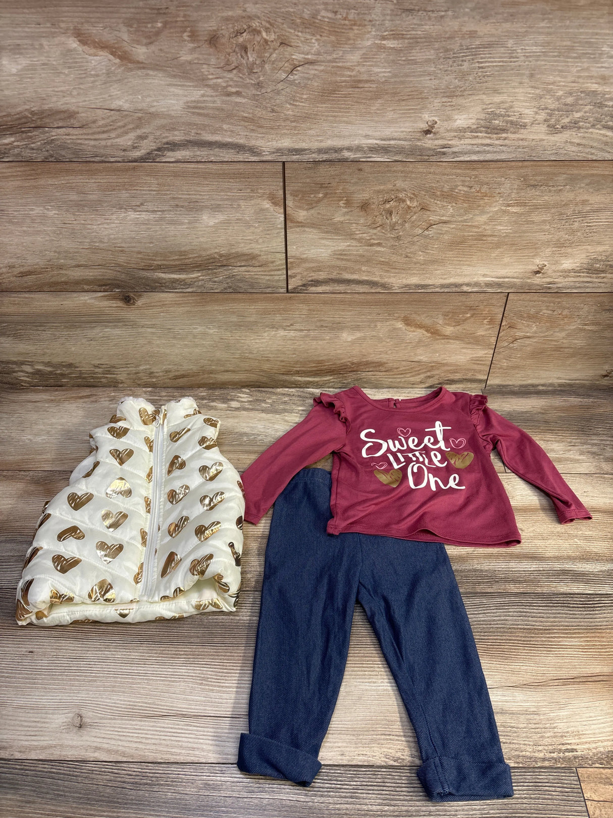 Flat lay of 3-piece toddler outfit: cream puffer vest with gold hearts, pink shirt reading ‘Sweet Little One’, blue jeggings