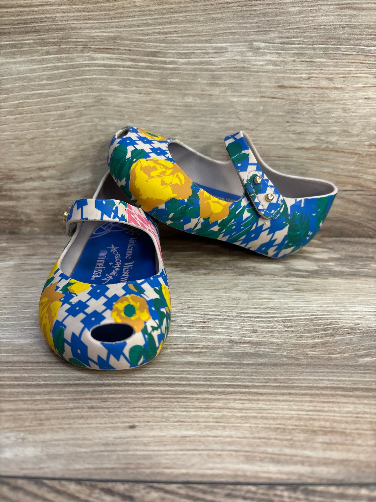 Front three-quarter view of Vivienne Westwood x Melissa Mini Ultra girls’ Mary Jane shoe, size 6C, showing blue, yellow and green floral print and peep-toe on woodgrain background