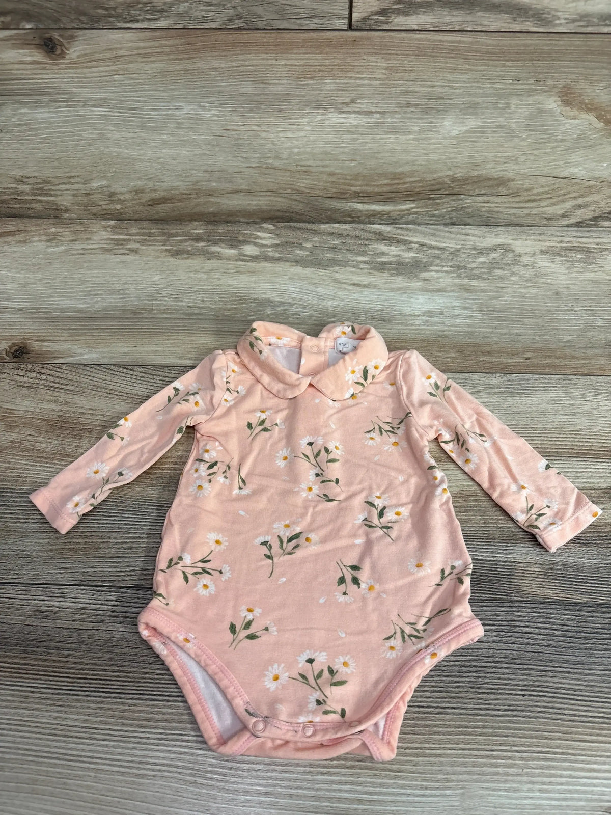 Flat lay of pink Angel Dear Peter Pan collar bodysuit showing daisy print, long sleeves, and snap crotch on wood background
