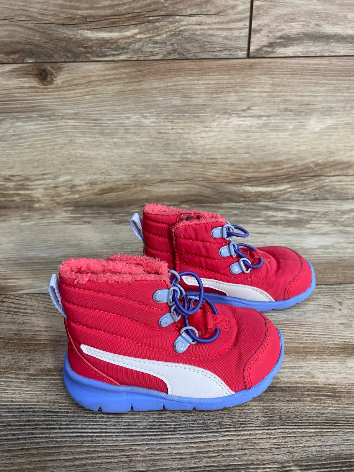 Side view of Puma Bao 3 AC toddler snow boots in pink showing blue soles, white Puma stripe and faux-fur lining on wood floor
